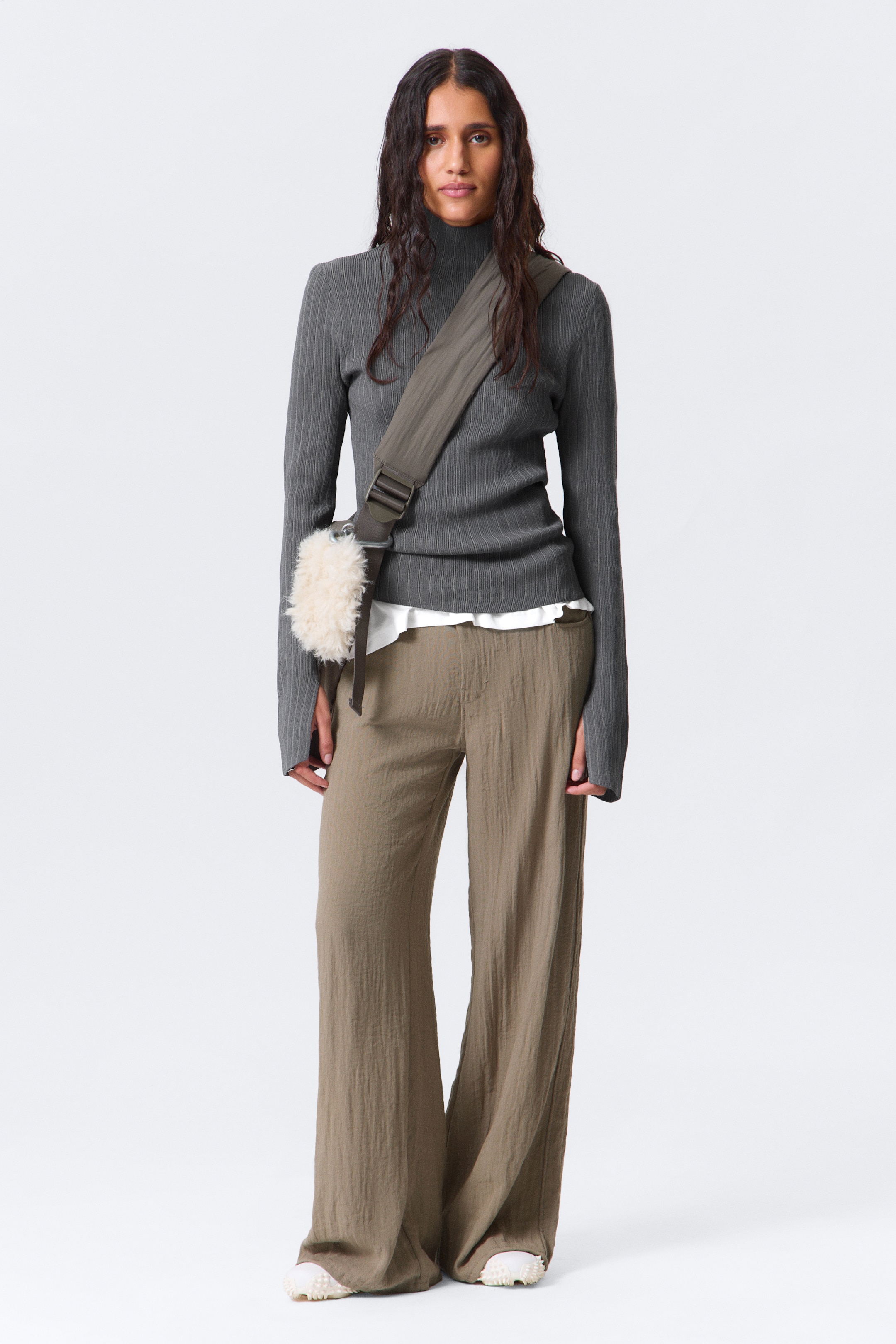 View larger image: A grey ribbed turtleneck sweater is being presented from a front-facing angle, featuring a high neck and vertical ribbing throughout its long sleeves and body. It is being layered over a white garment, with its hem visible, and styled with olive green wide-leg pants, which are showing a crinkled texture.