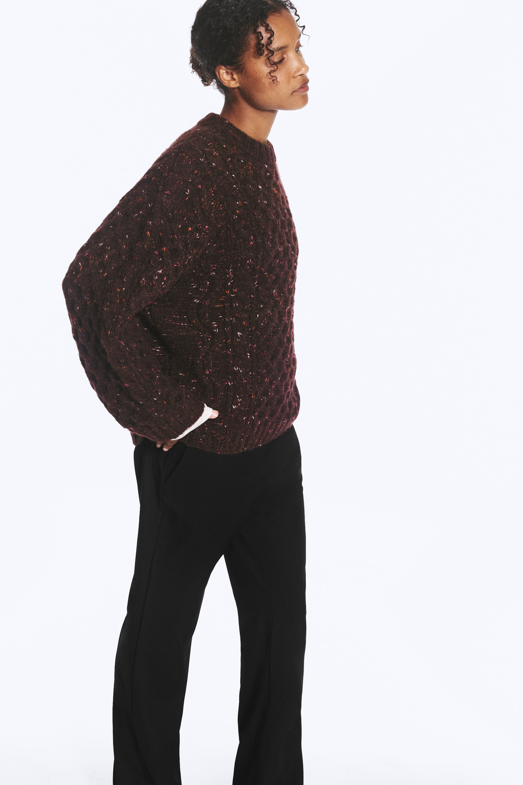 Größeres Bild ansehen: Ein dunkelbrauner, melierter Zopfpullover mit strukturierter Oberfläche, weiten Ärmeln und Rippbündchen wird vom Model getragen. Der Pullover hat eine lockere Passform und wird mit einer schwarzen Hose kombiniert.