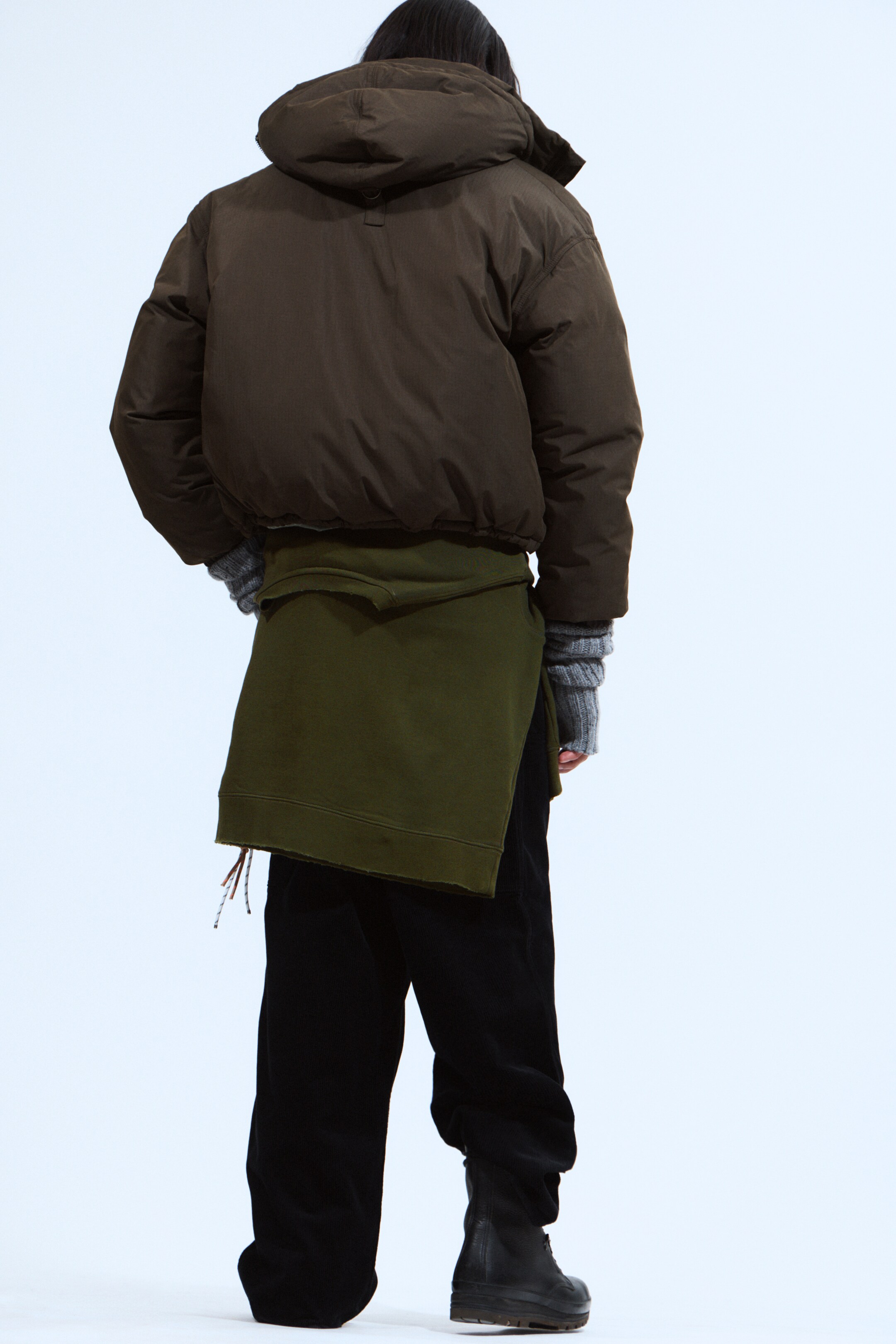 Agrandir l'image: Une doudoune marron foncé est montrée de dos, légèrement inclinée vers la droite, avec une coupe volumineuse et raccourcie et une capuche intégrée. Des gants gris côtelés sans doigts s'étendent de ses manches, et un vêtement vert enveloppant avec des bords bruts est superposé en dessous.
