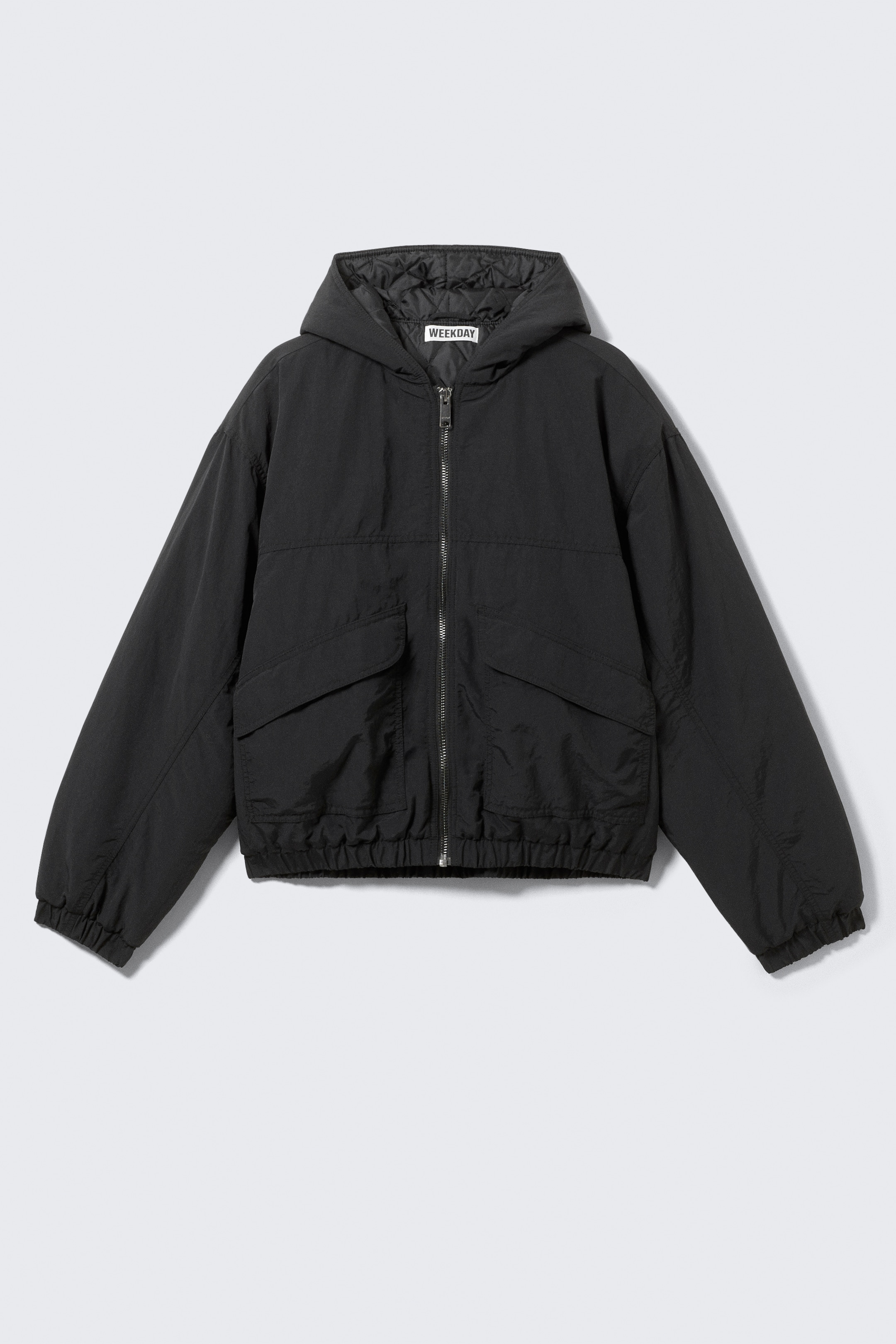 Visualizza immagine più grande: Una giacca bomber in nylon nero con cappuccio è mostrata dalla parte anteriore, mostrando una vestibilità rilassata. Presenta un cappuccio spazioso con fodera trapuntata visibile, una robusta chiusura frontale con zip, due tasche frontali con patta in velcro, polsini elasticizzati e un orlo elasticizzato.