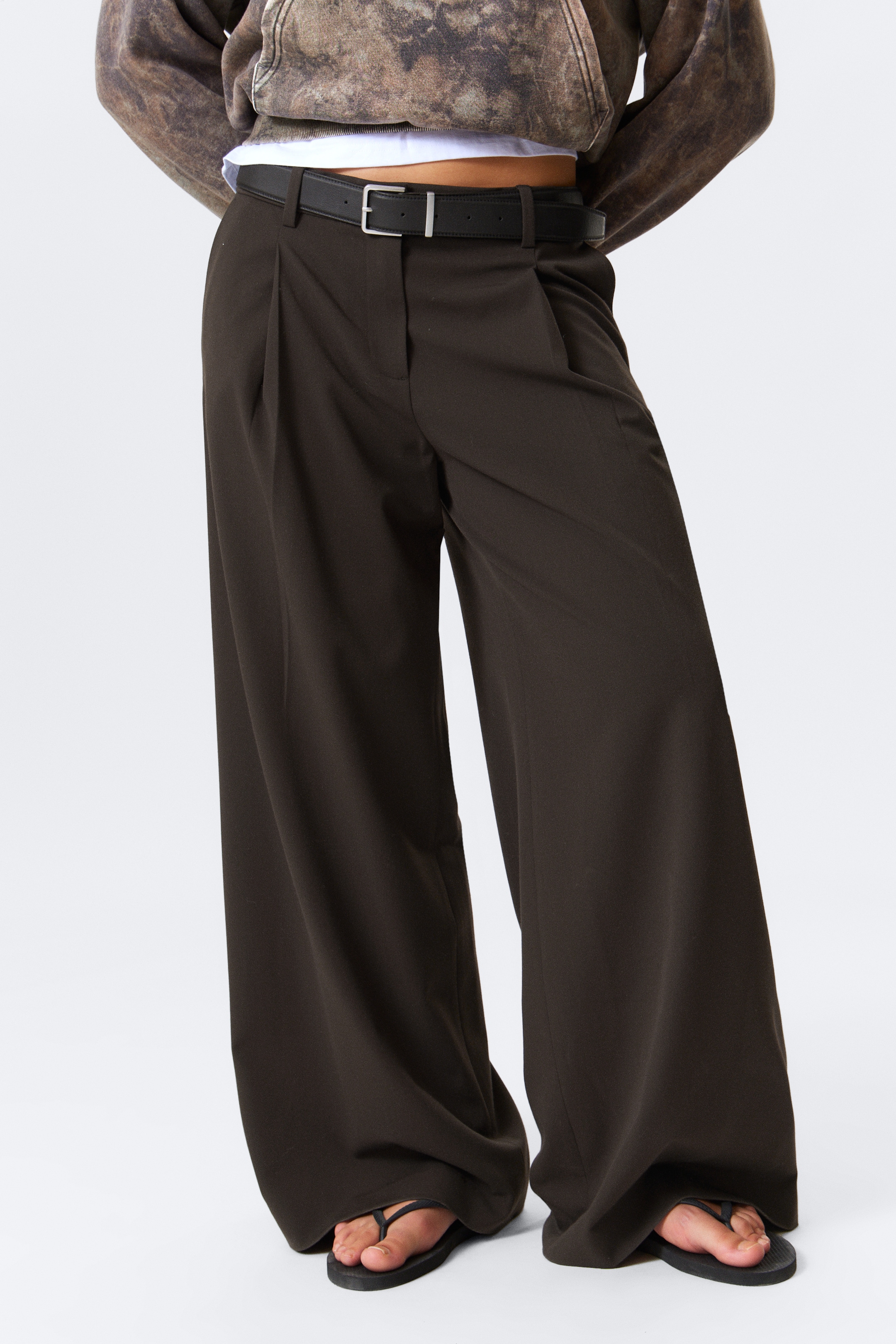 Agrandir l'image: Un pantalon large de couleur marron foncé est présenté de face, s'étendant jusqu'au sol et s'accumulant légèrement sur les pieds. Ils présentent des plis à la taille avant et sont maintenus par une ceinture noire.