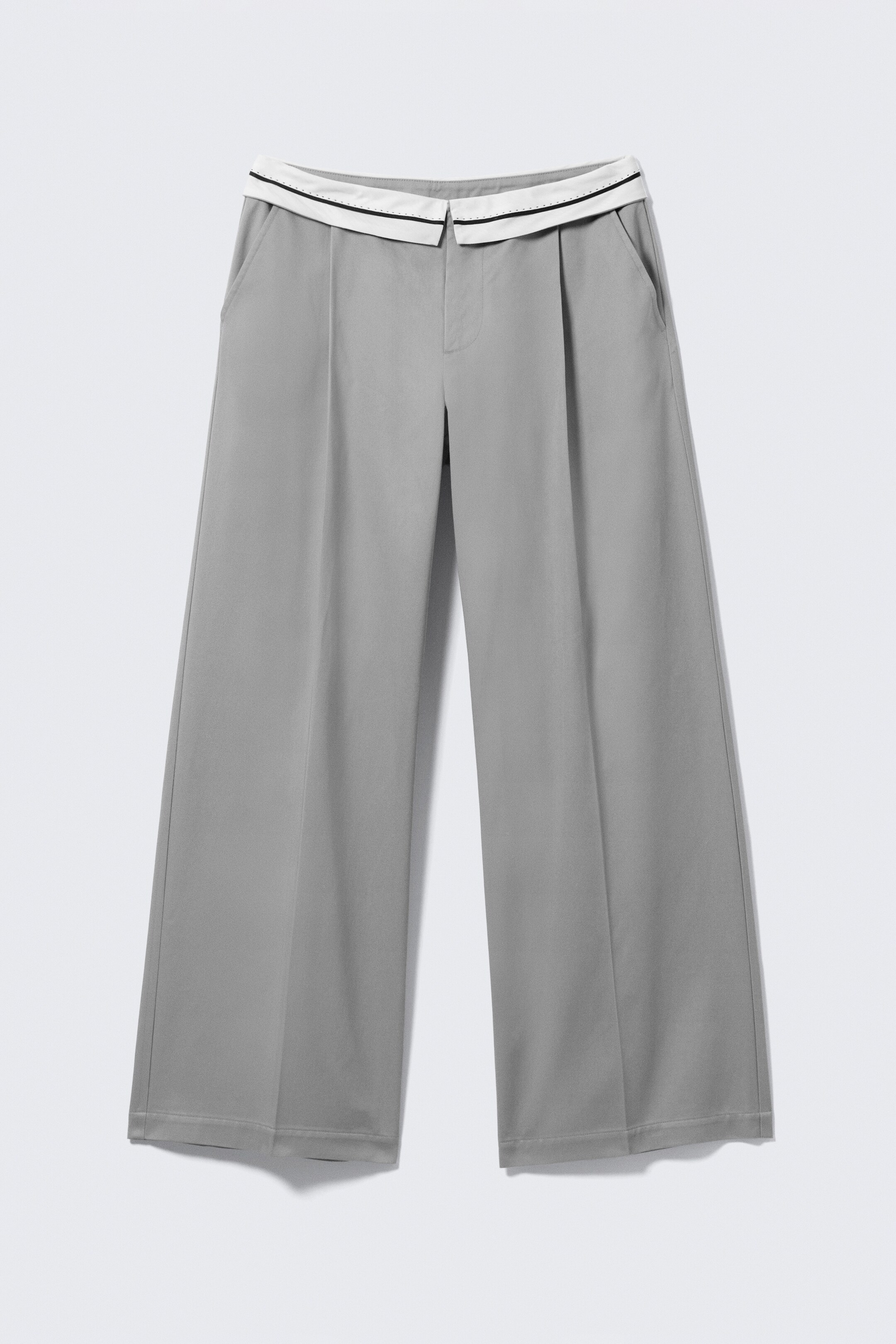 Grotere afbeelding bekijken: Een paar grijze katoenen twill lage taille broeken presenteert FRONT, met een gestructureerd silhouet met rechte pijpen. De broek toont een contrasterende omgevouwen tailleband in wit met een zwarte streep, geperste plooien langs de voorkant van elke pijp en schuingeplaatste zijzakken.