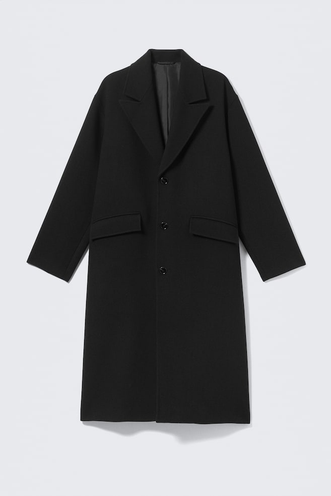 En lång, svart oversized kappa visas framifrån, med en avslappnad silhuett, slag med notcher och enkelknäppning framtill med tre knappar. Kappan är designad med två fickor med lock framtill, långa ärmar och en rak fåll med ren finish. - 1