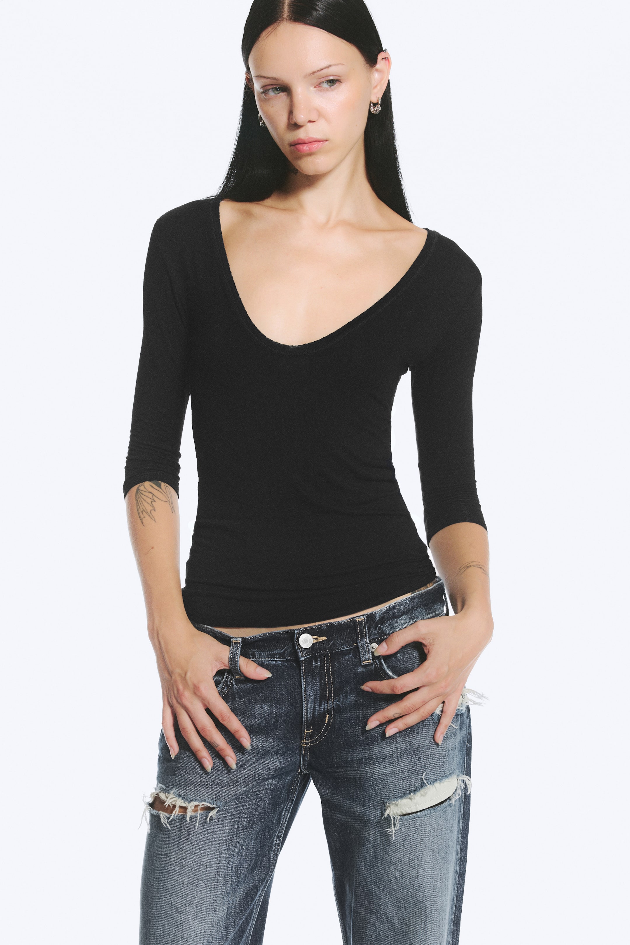 View larger image: A form-fitting black top featuring a deep scoop neckline and three-quarter-length sleeves is worn by the model. The top is styled with low-rise, distressed denim jeans, with the model resting their hands near the front pockets.