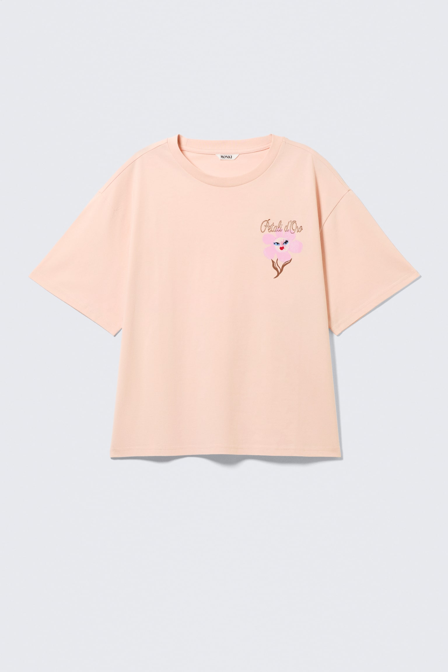 Ein helles Pfirsich-T-Shirt präsentiert sich mit einer lockeren, kastigen Passform, überschnittenen Schultern und einem Rundhalsausschnitt von der VORDERSEITE. Ein stilisiertes Blumenmotiv mit rosa Blütenblättern, einem blauen Zentrum und einem braunen Stiel sowie dem Text 'Pétali d'Oro' ist auf der linken Brustseite aufgedruckt und wird mit geraden Säumen abgeschlossen. - 1