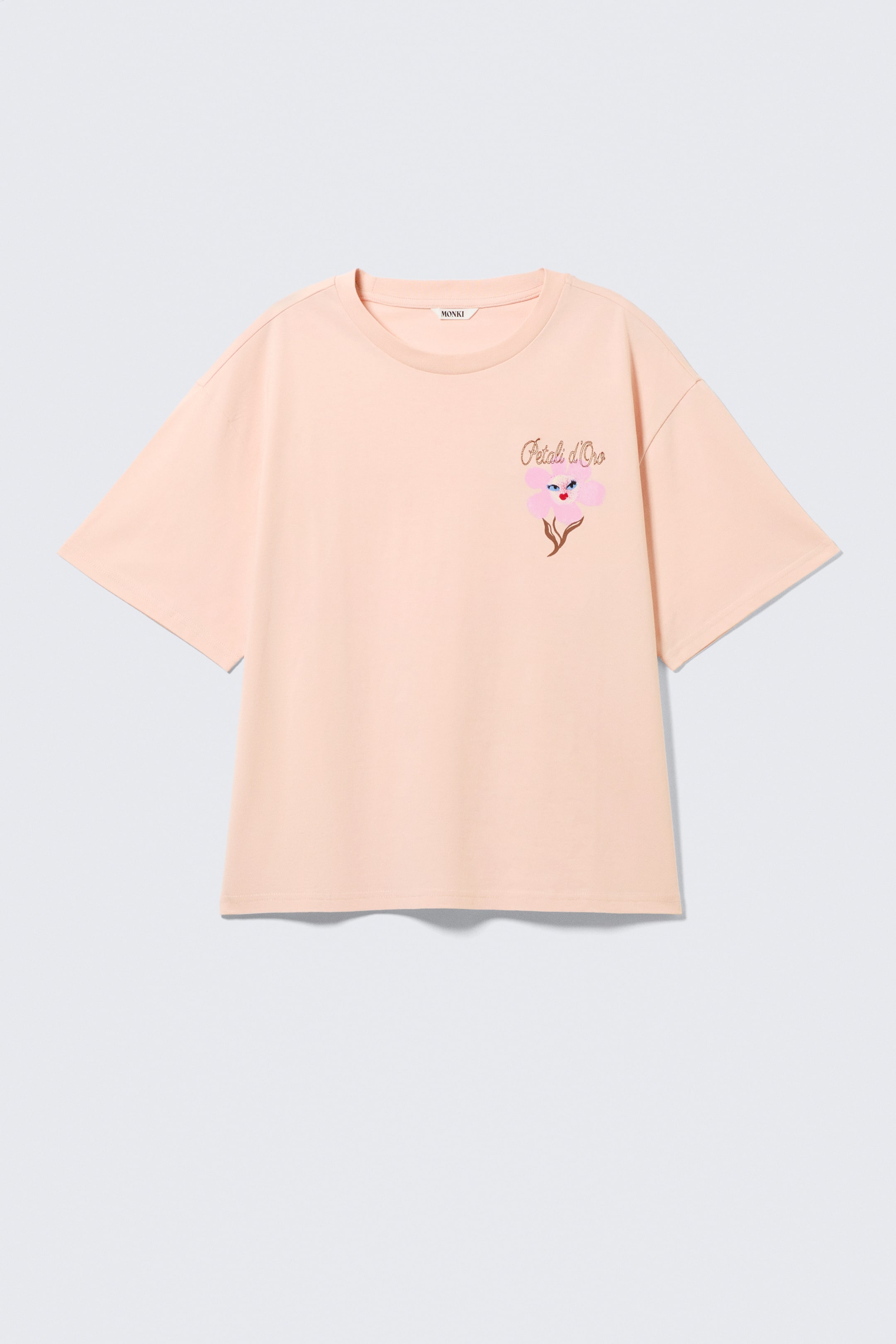 Visualizza immagine più grande: Una t-shirt color pesca chiaro presenta una vestibilità ampia e squadrata con spalle cadenti e scollo a girocollo nella parte anteriore. Un grafico floreale stilizzato, con petali rosa, un centro blu e uno stelo marrone, con il testo 'Pétali d'Oro' è stampato sul petto sinistro anteriore, e si completa con orli dritti.