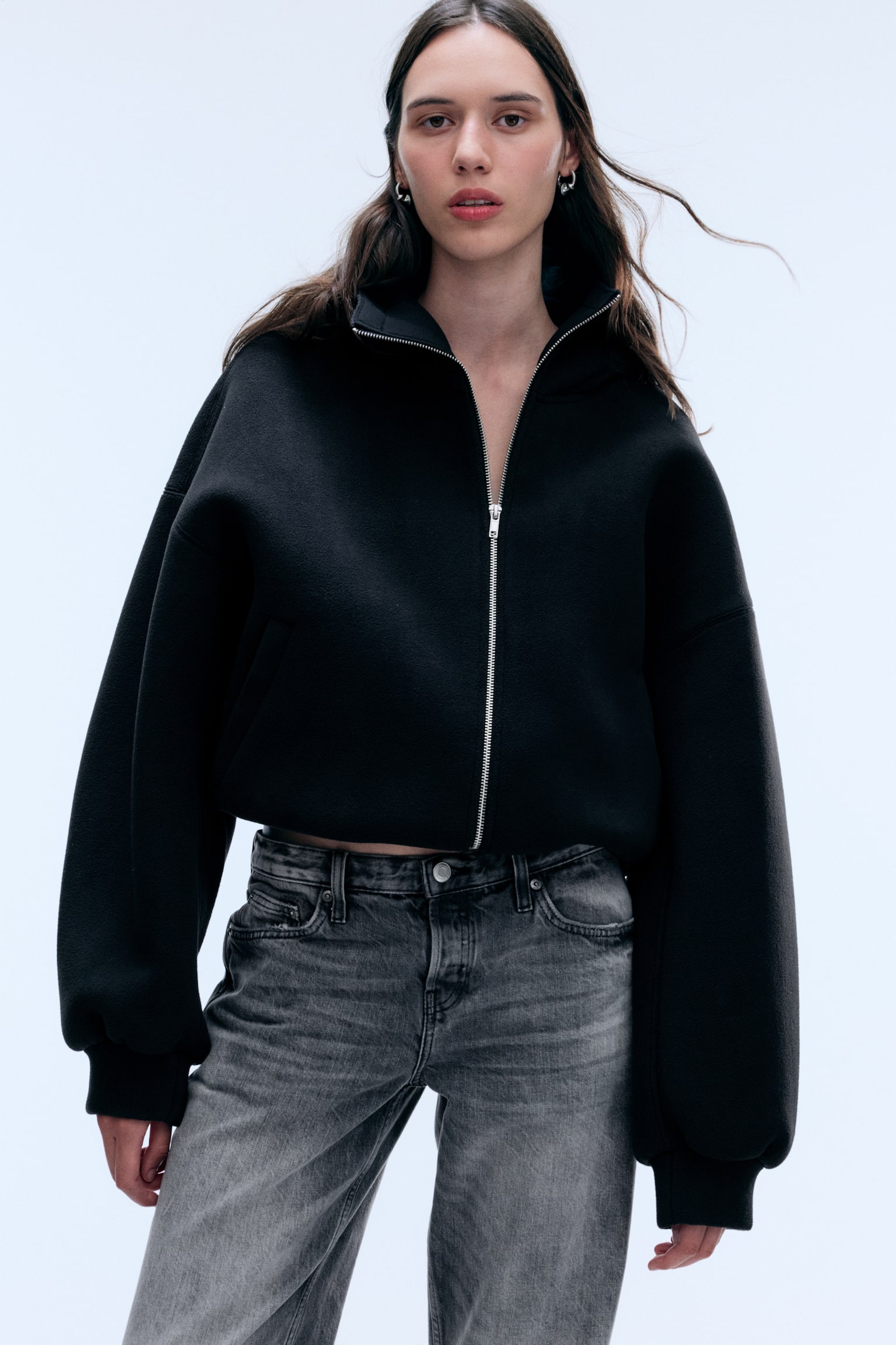 Model má na sebe oversized Äiernu zipsovú bundu so Å¡truktúrovanou textúrou, doplnenú sivými vypranými džÃnsami. Bunda má spustené ramená, objemné dlhé rukávy s rebrovanými manžetami a strieborný predný zips, ktorý je ÄiastoÄne zapnutý do vysokého goliera. - 1