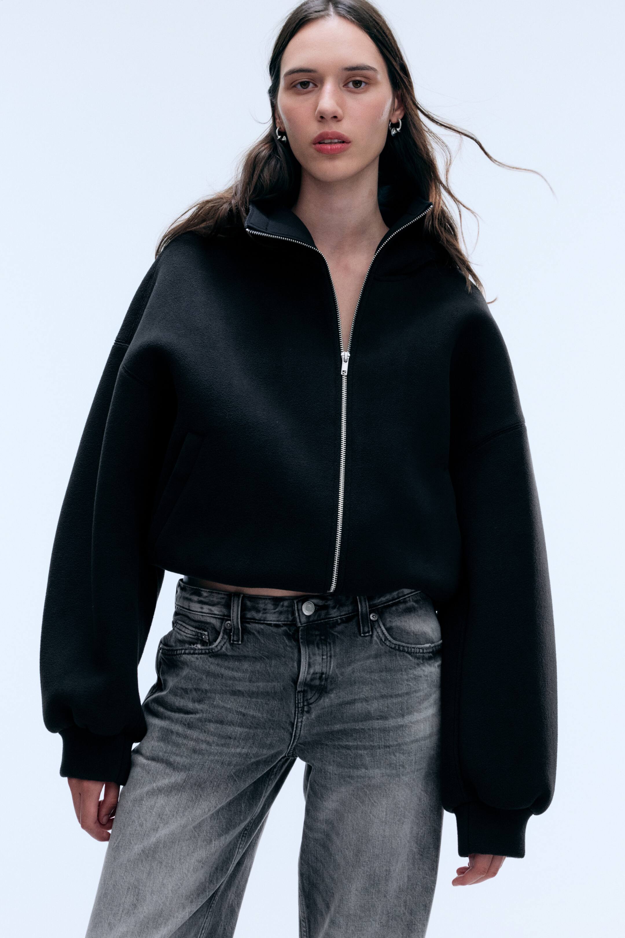 Visa större bild: En svart oversized jacka med dragkedja och strukturerad textur bärs av modellen, tillsammans med grå, tvättade jeans. Jackan har nedhasade axlar, voluminösa långa ärmar med ribbade muddar och en silverfärgad dragkedja framtill som är delvis stängd för att bilda en hög krage.