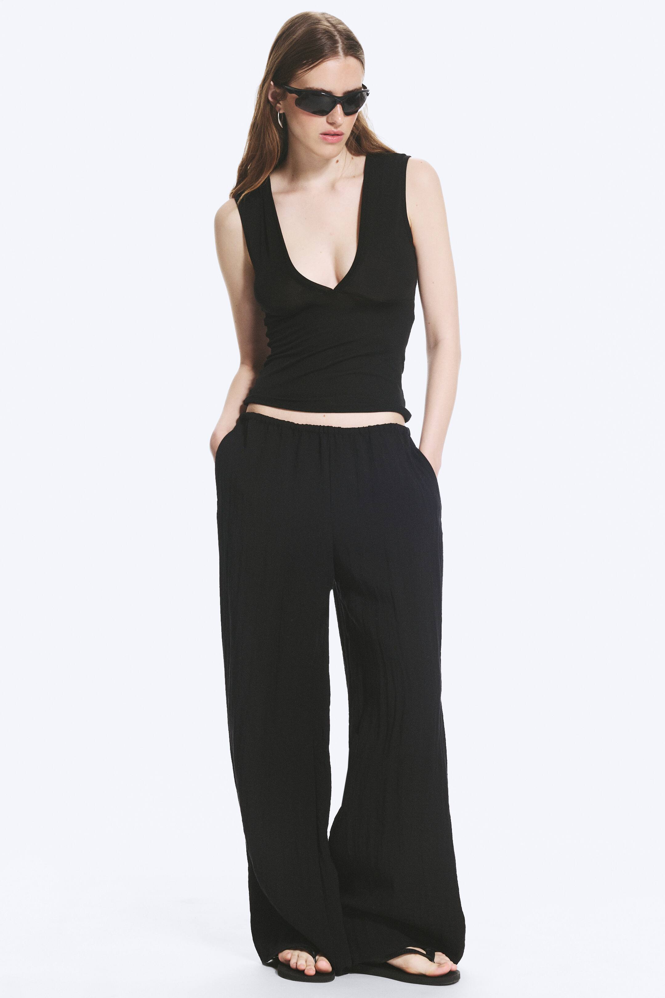 Ver imagen más grande: Pantalones negros, confeccionados con un tejido ligero arrugado y un acabado texturizado, están siendo llevados por la modelo. Cuentan con una cintura elástica baja y piernas anchas de largo completo. La modelo está de pie con las manos en los bolsillos, combinándolos con un top negro sin mangas y chanclas negras.