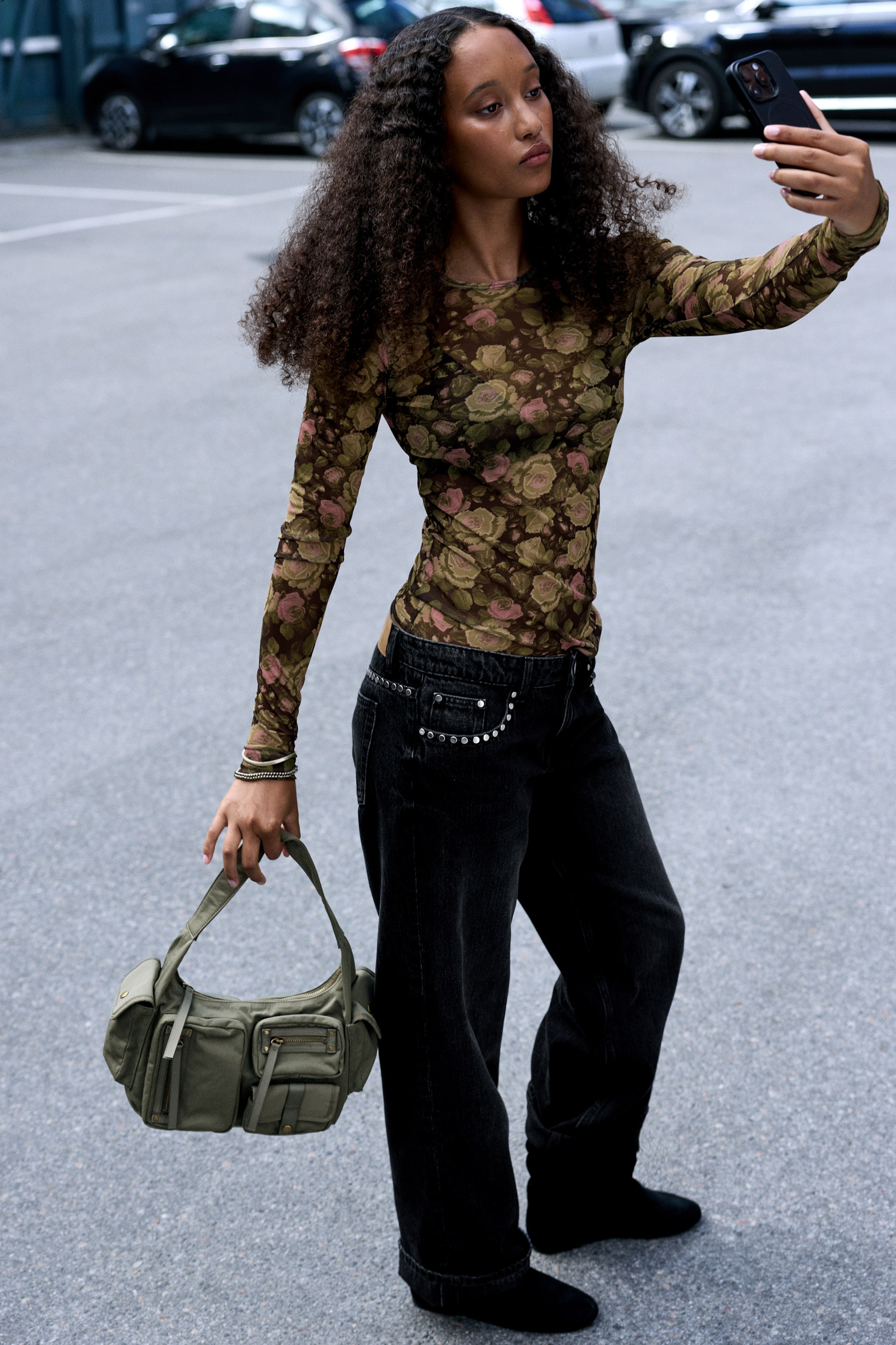 Visualizza immagine più grande: Un top stampato a maniche lunghe dalla vestibilità slim, caratterizzato da un motivo floreale verde, marrone e rosa, indossato dalla modella. La modella abbina il top girocollo a jeans scuri a gamba larga, tenendo uno smartphone in una mano e portando una borsa verde nell'altra.