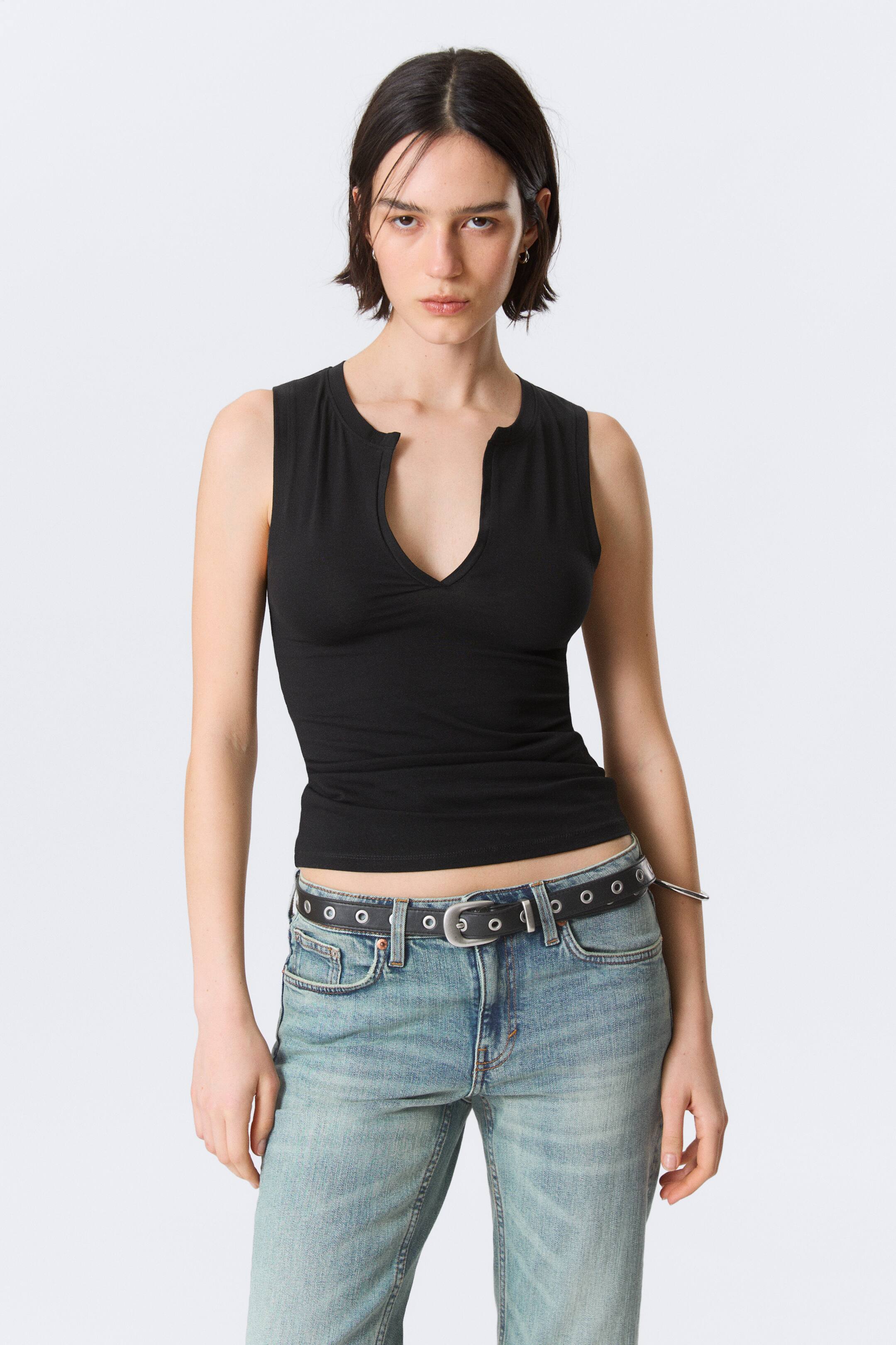Ingrandisci l'immagine: La modella indossa un top nero senza maniche con scollo a V incavato e silhouette aderente, abbinato a jeans chiari e una cintura nera con borchie.