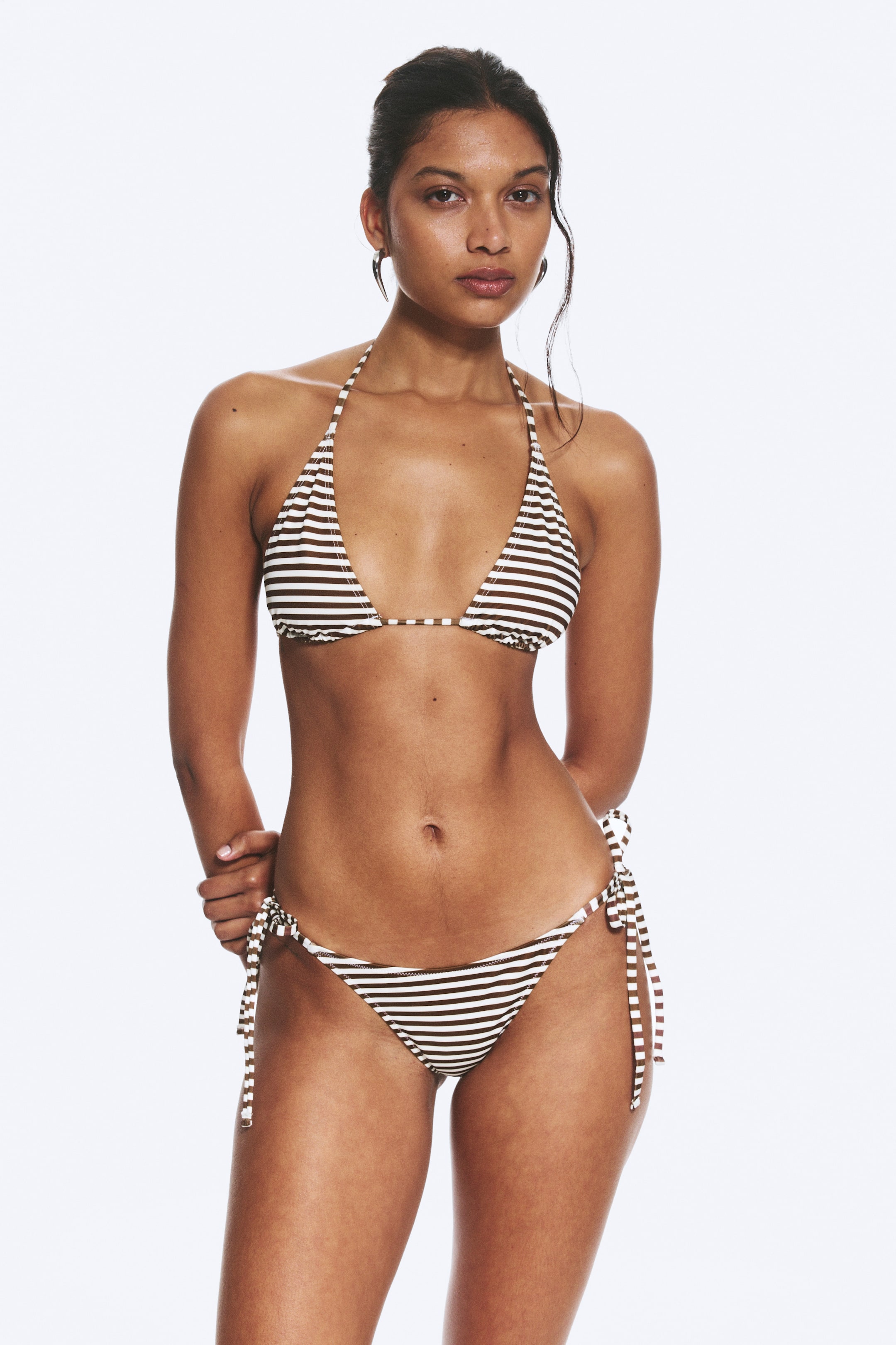 Größeres Bild ansehen: Ein zweiteiliger Bikini mit braunen und weißen horizontalen Streifen wird von dem Model getragen. Das Triangel-Oberteil verfügt über Nacken- und Rückenschnürungen, und das brasilianische Unterteil hat schmale Bänder, die an den Hüften gebunden werden.