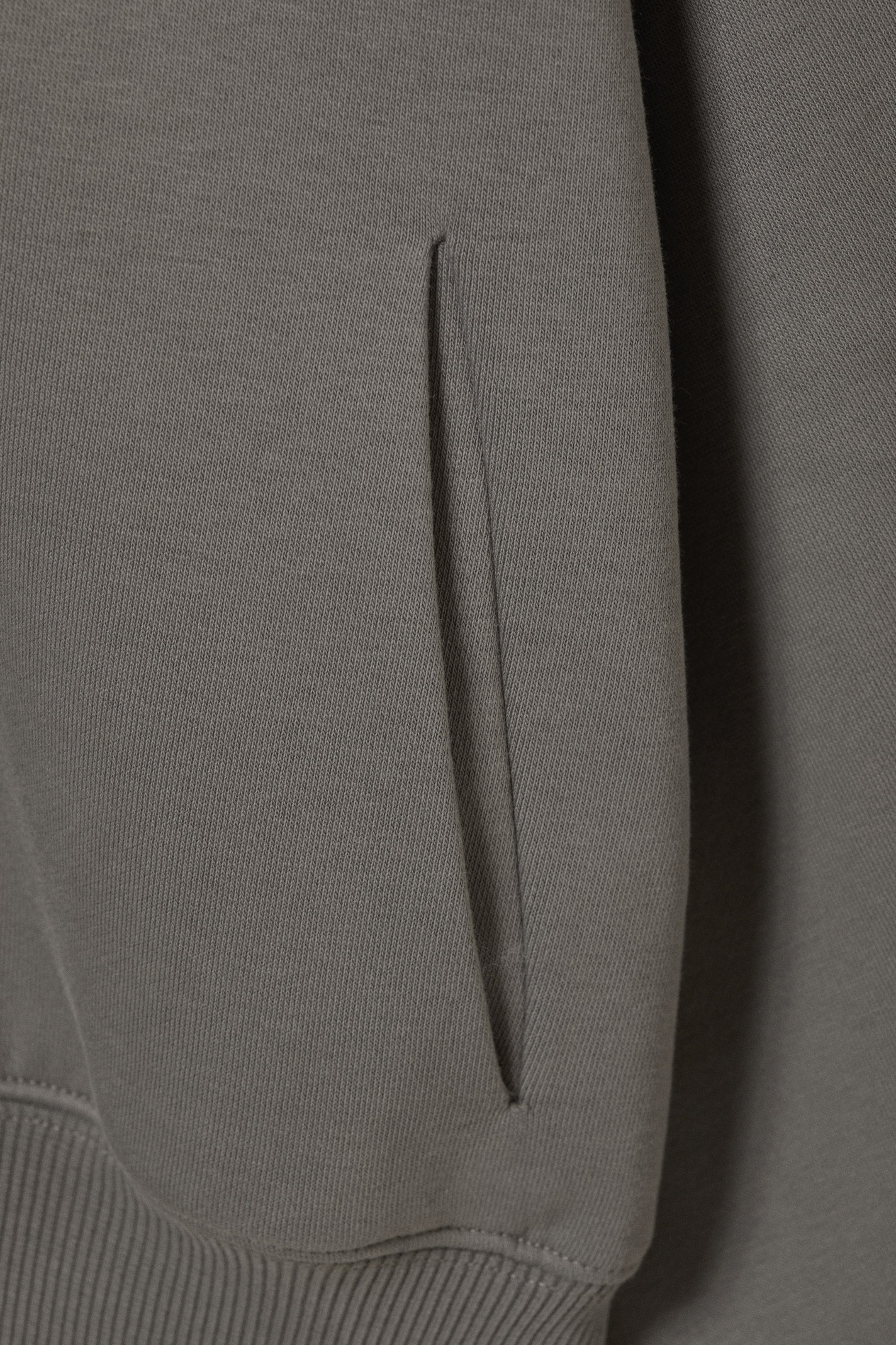 Agrandir l'image: Un sweat à capuche en molleton de coton gris est présenté en gros plan, mettant en valeur une poche discrète le long de la couture latérale. Un ourlet large et côtelé termine le bas du vêtement, avec des surpiqûres ton sur ton visibles autour des zones de la poche et de l'ourlet.