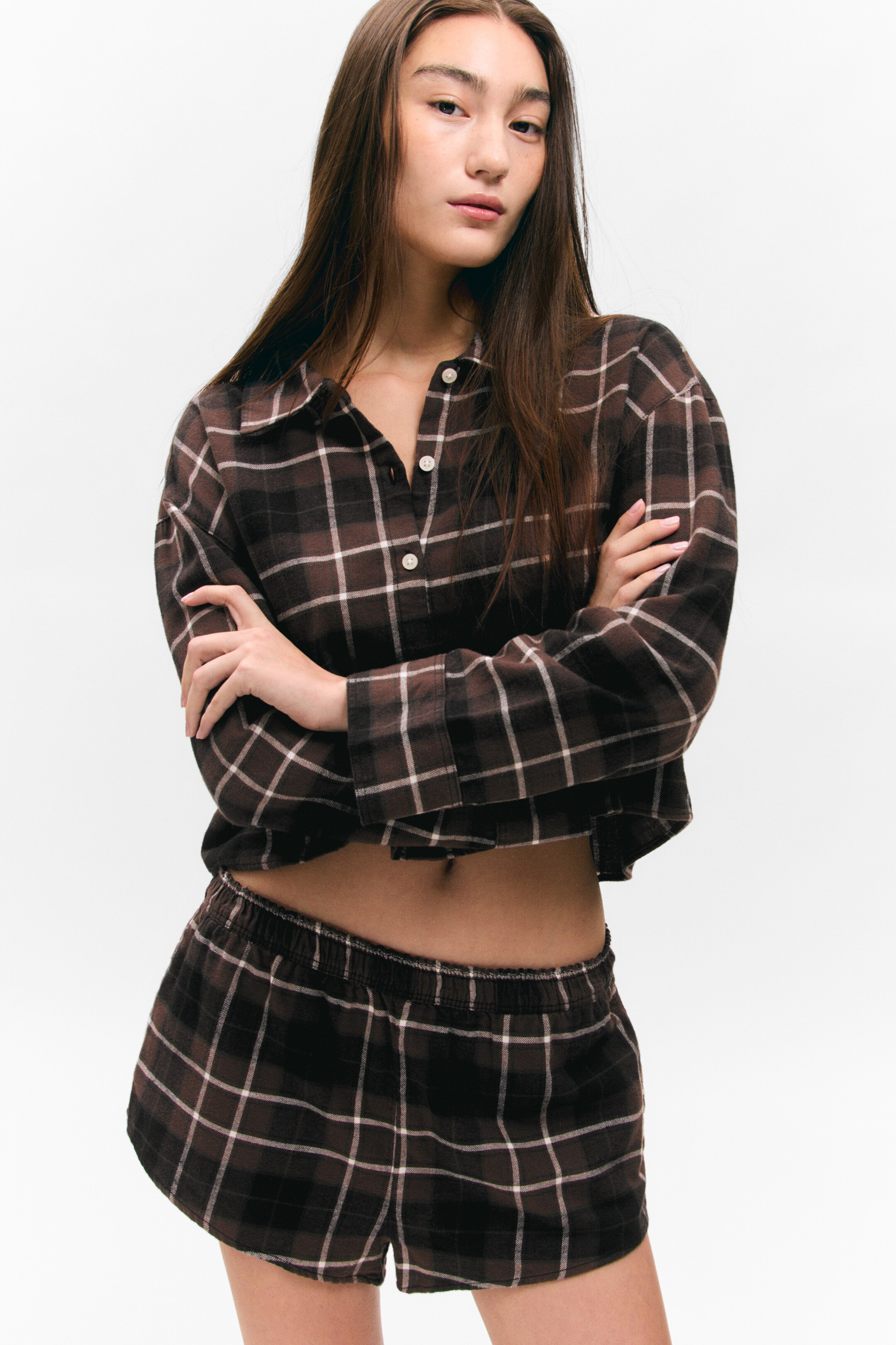 Ingrandisci l'immagine: Un set da notte a quadri marroni e bianchi viene presentato frontalmente, caratterizzato da una camicia corta a maniche lunghe e shorts coordinati. La camicia è progettata con un colletto e un'abbottonatura parziale sul davanti, mentre gli shorts presentano una cintura elastica.