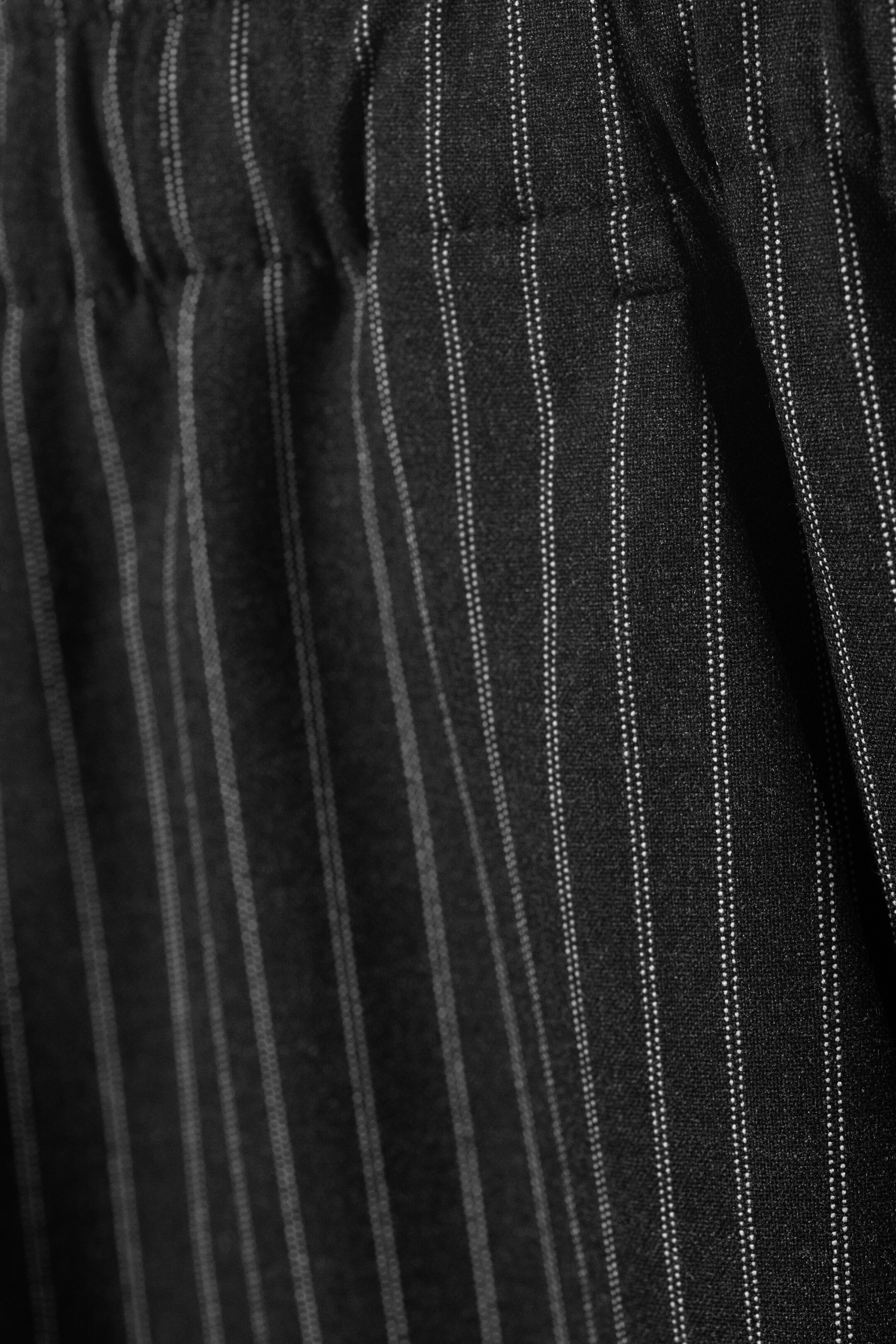 Agrandir l'image: Un pantalon de costume apparaît en gros plan, montrant un tissu sombre avec de fines rayures verticales blanches et une taille froncée et élastiquée.
