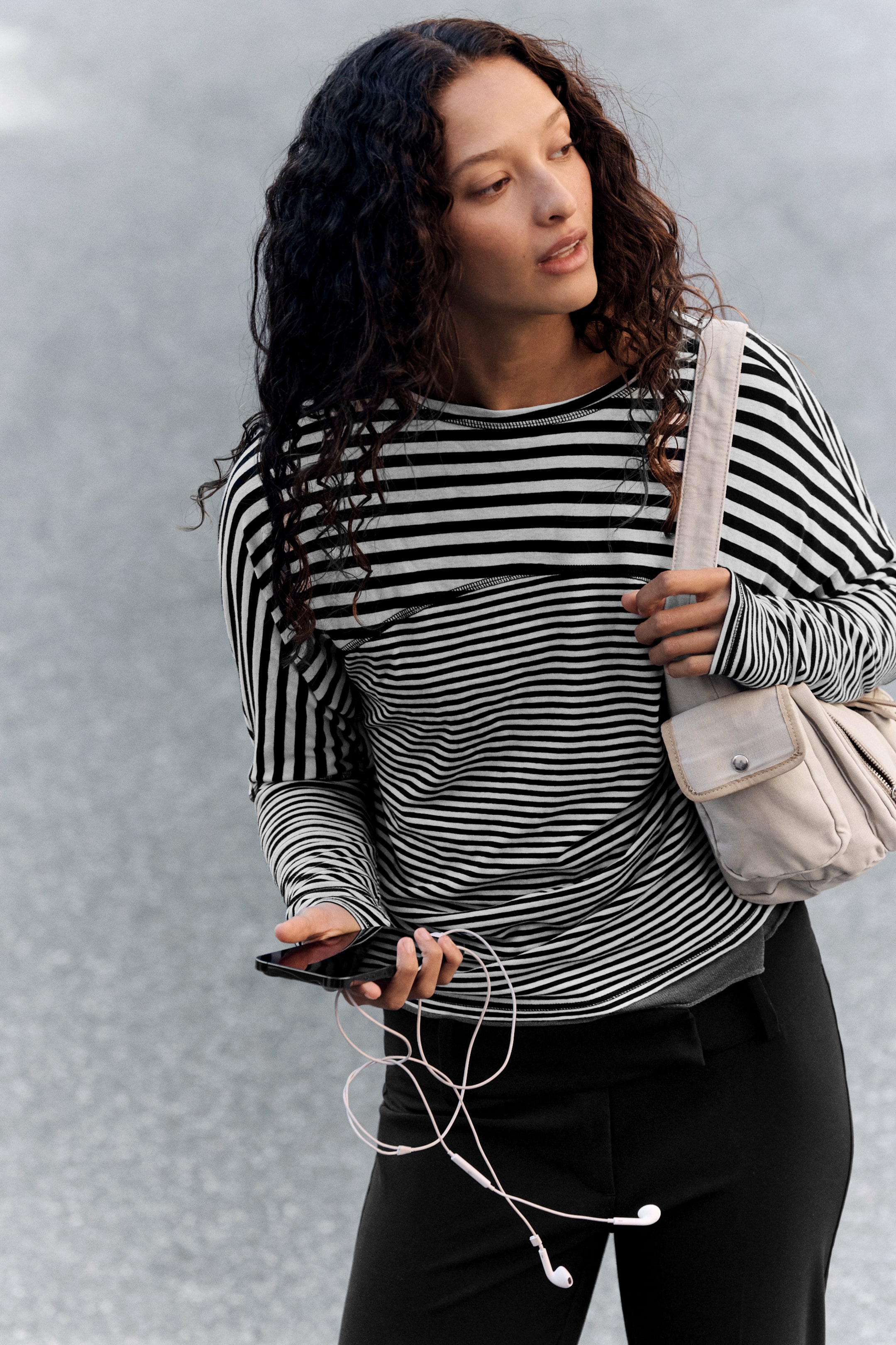 Grotere afbeelding bekijken: Een zwart-wit gestreept T-shirt met lange mouwen, batwingmouwen en zichtbare whipstitch-naden langs de gebogen paneelnaden wordt gedragen door het model. Het model combineert het bovenstuk met een zwarte broek en een lichtgekleurde schoudertas, terwijl hij of zij een smartphone vasthoudt.