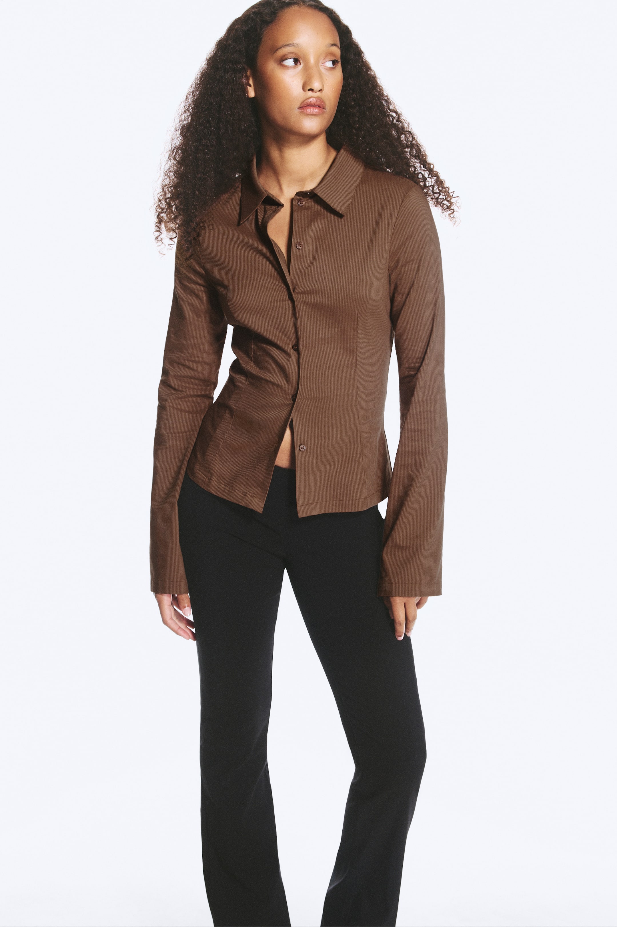 Agrandir l'image: Une chemise marron ajustée, dotée d’un col classique, d’une fermeture boutonnée sur le devant et de manches extra-longues, est portée par le mannequin. La chemise est associée à un pantalon noir.