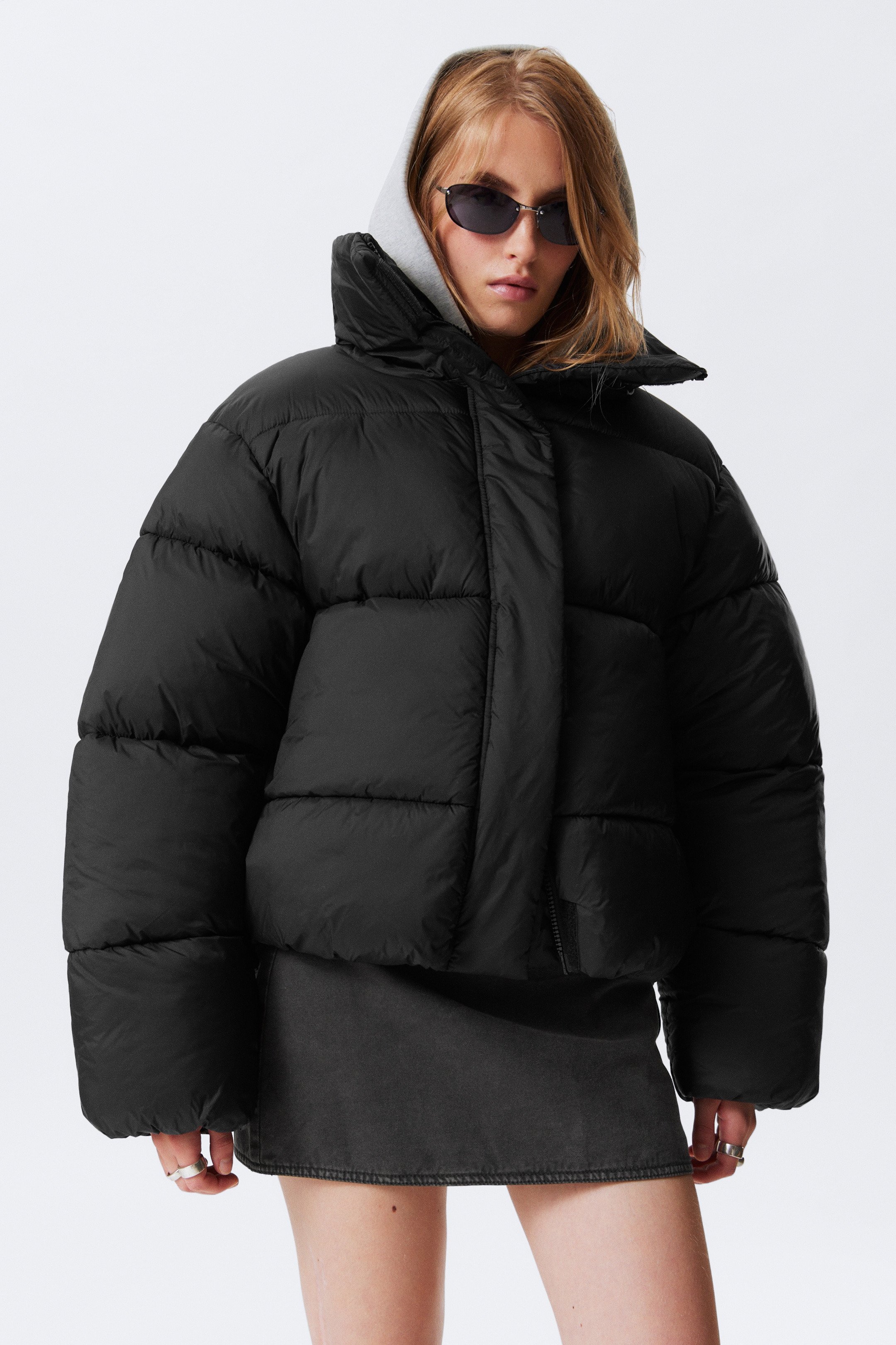 Ingrandisci l'immagine: Una giacca nera oversize corta e imbottita è presentata dal davanti, caratterizzata da un colletto alto, trapuntatura orizzontale e una chiusura frontale nascosta. Una felpa con cappuccio grigio chiaro è visibile sotto il colletto della giacca, indicando la stratificazione.
