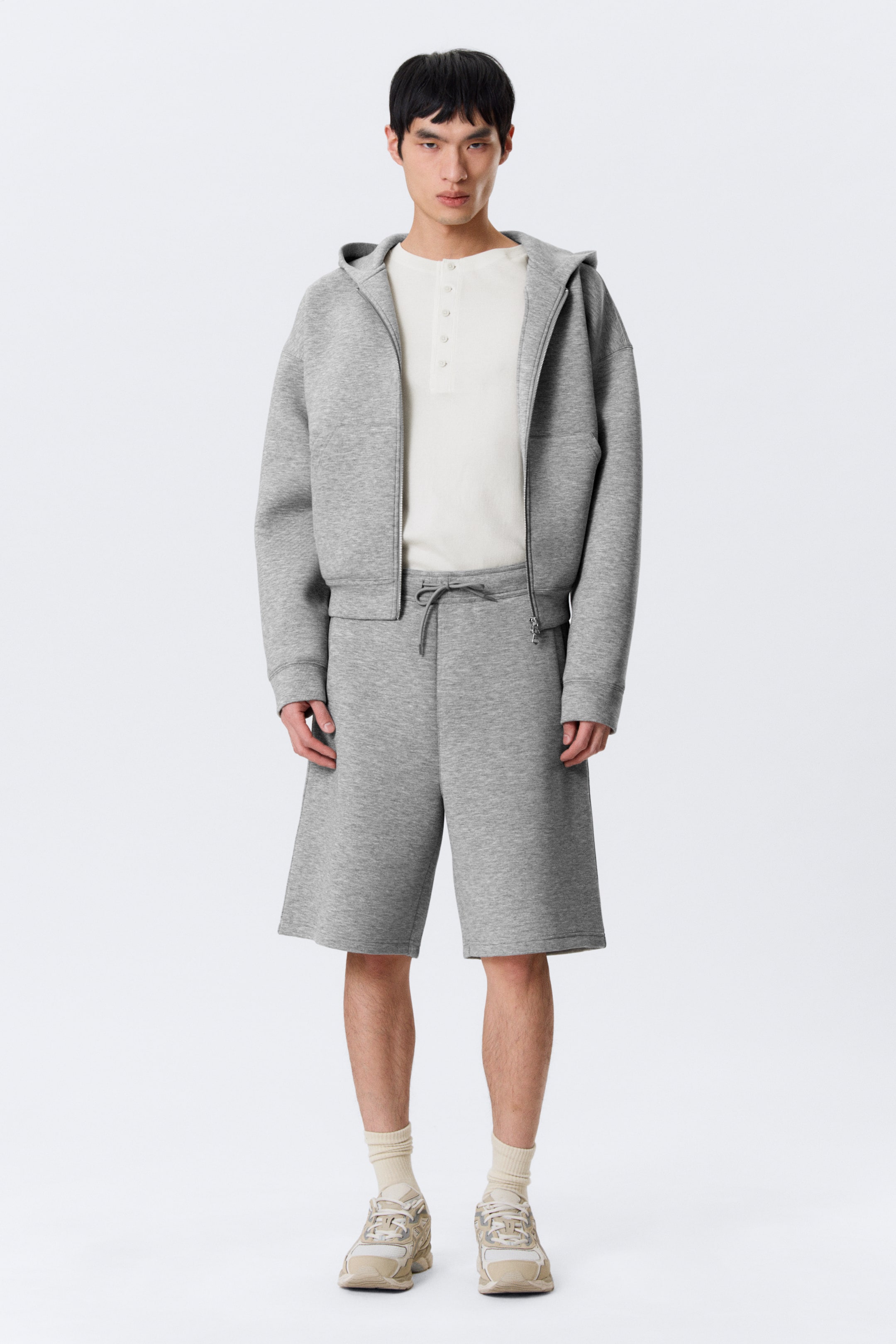 Grotere afbeelding bekijken: Een grijze gestructureerde gebreide loungewear set wordt frontaal gepresenteerd, met een hoodie met rits en capuchon en bijpassende knielange shorts met een trekkoord in de taille. De hoodie wordt open gedragen, waardoor een crèmekleurig Henley-shirt met vier knopen eronder zichtbaar is.