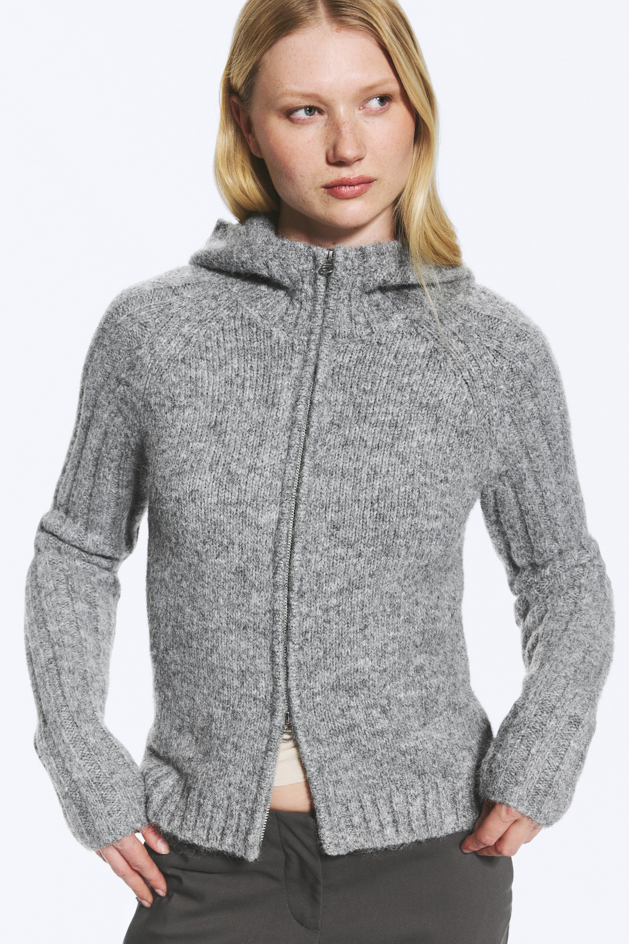 Ver imagen más grande: Una sudadera con capucha de punto gris, con mangas acanaladas y capucha, llevada por la modelo. El cierre de cremallera completa está parcialmente abierto, dejando ver una prenda de color claro debajo.