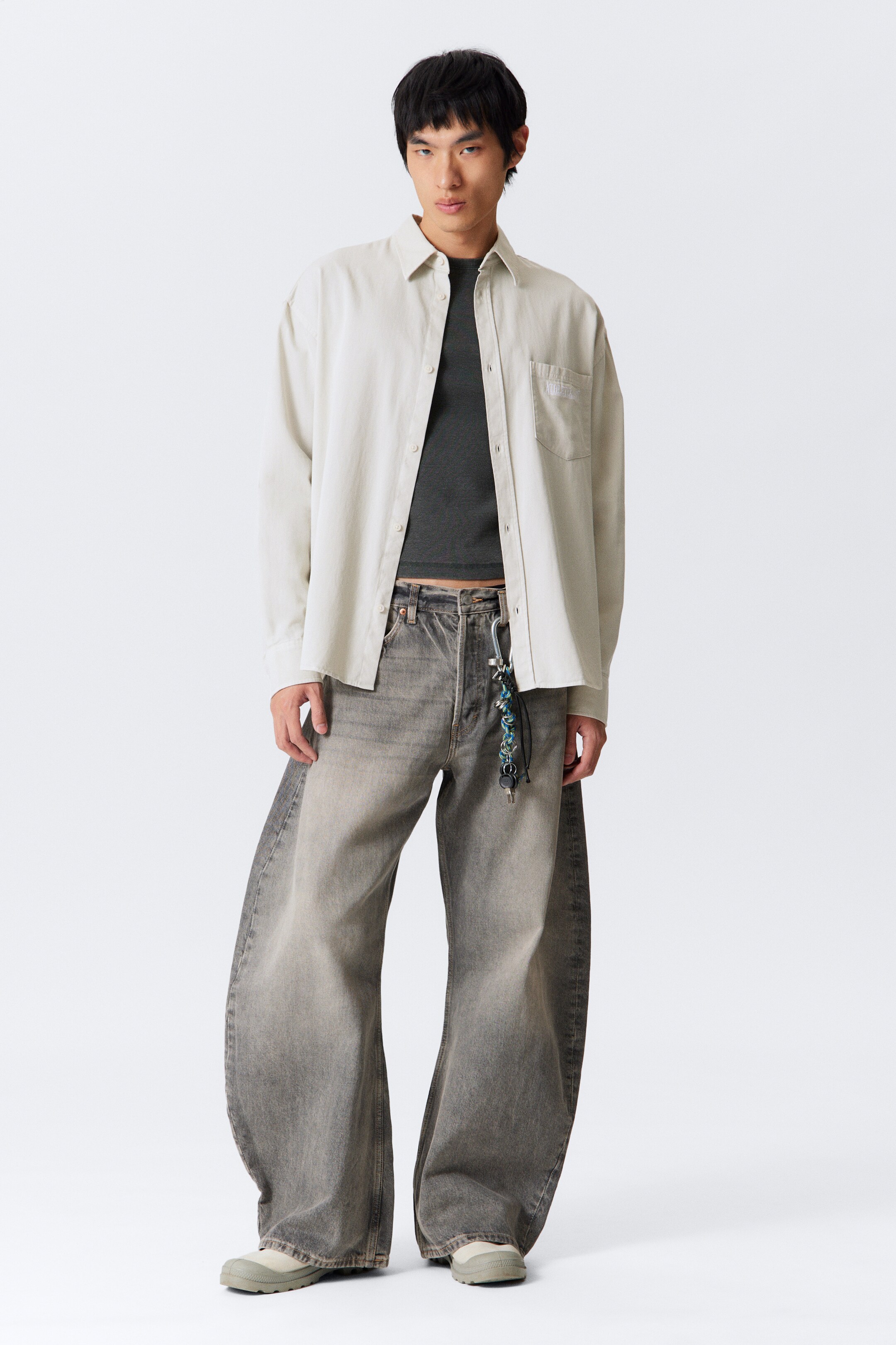 Visa större bild: En outfit visas upp, med en ljusbeige oversized skjorta med knappar och en bröstficka med synlig broderi, som bärs uppknäppt över en mörkgrå kortärmad topp. Skjortan är kombinerad med urtvättade gråa byxor med vida ben, som samlas över ljusgråa klumpiga skor.
