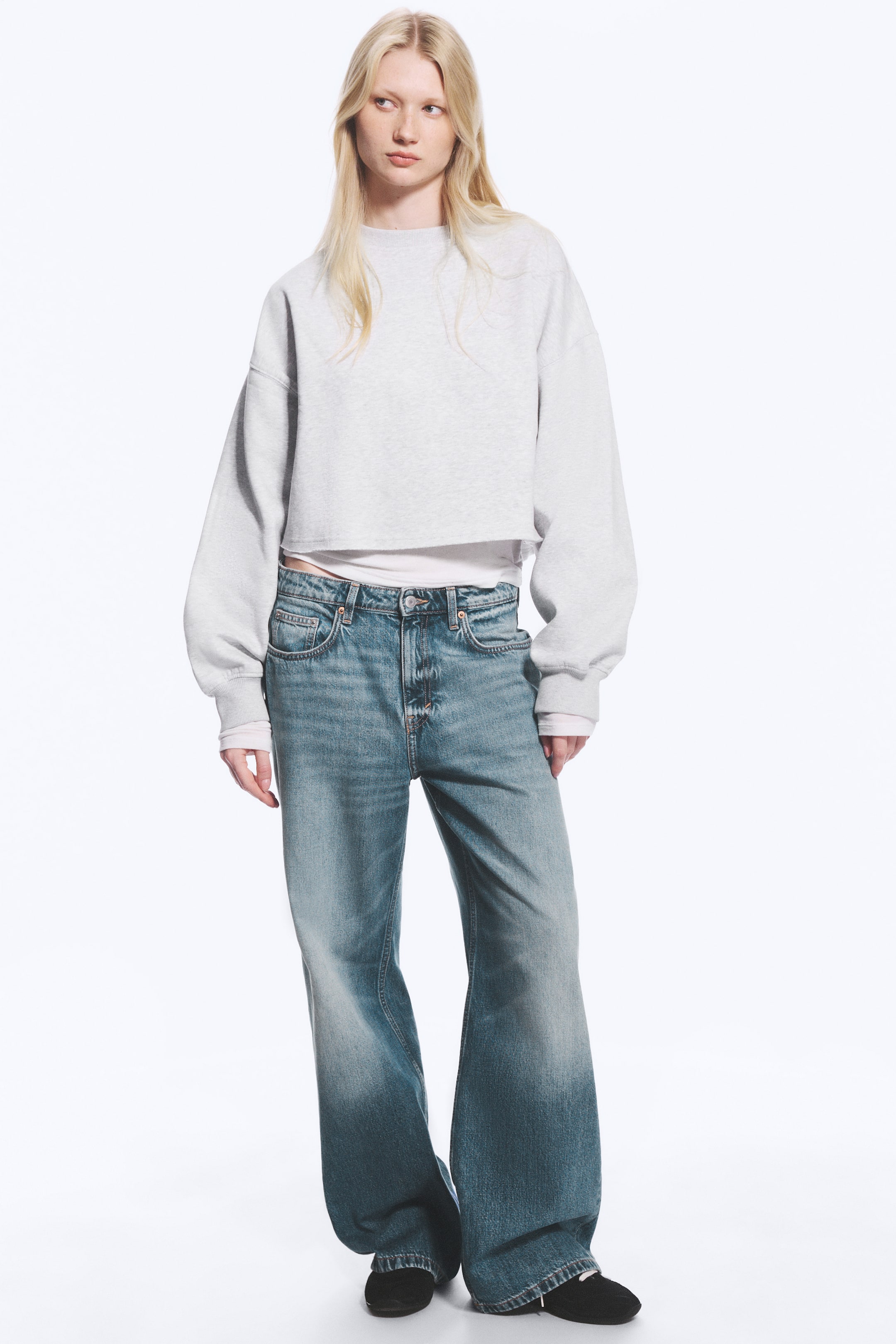 Agrandir l'image: Le mannequin porte une paire de jeans bleus à taille basse, à la coupe ample et à jambe droite. Le jean présente un délavage usé, un design classique à cinq poches et une braguette zippée, et il est associé à un sweat-shirt gris court et à des chaussures noires.