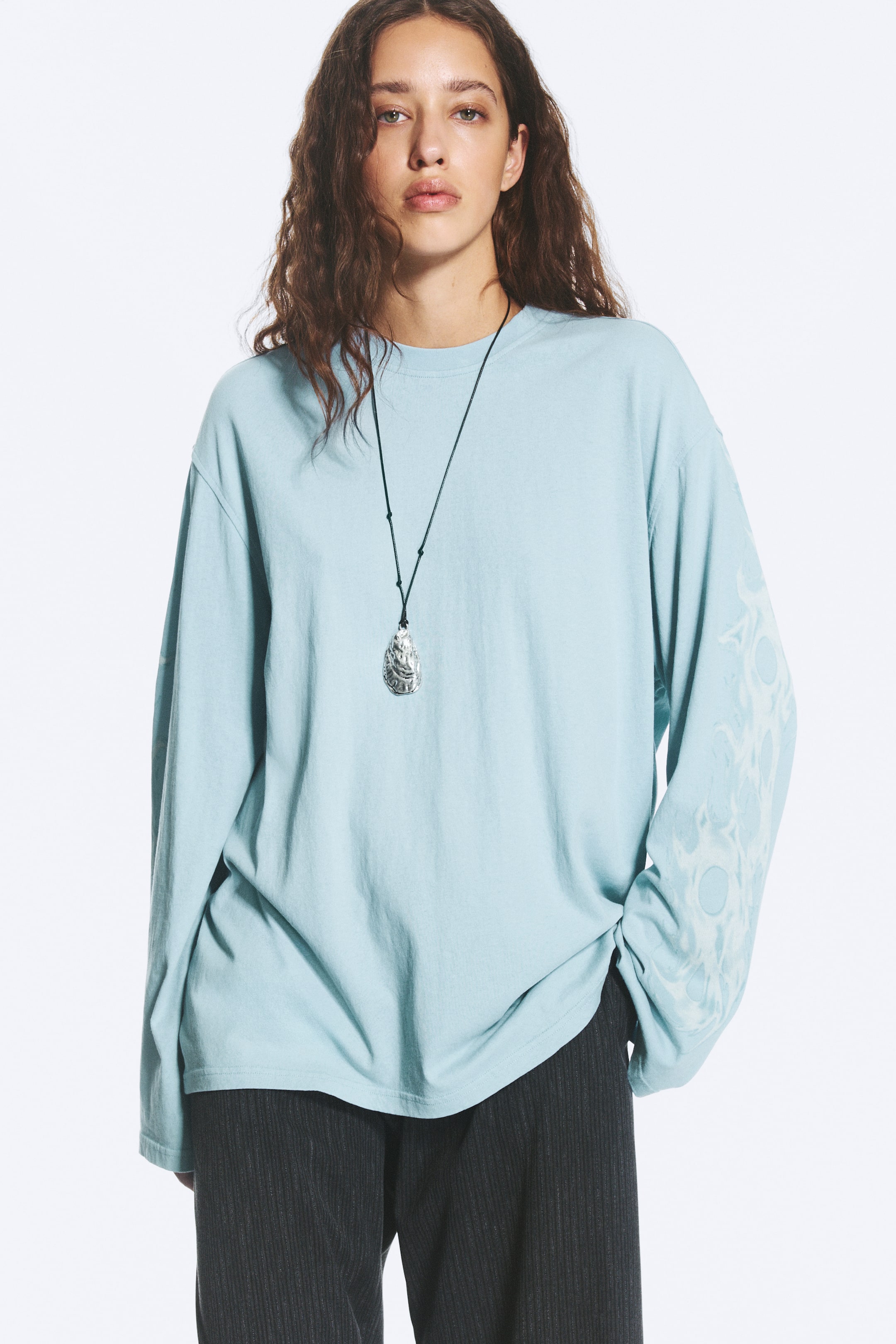 Ver imagen más grande: La modelo lleva puesta una camiseta de manga larga oversize de color azul claro. Presenta cuello redondo y un estampado de remolinos en tono claro en las mangas. La camiseta está parcialmente metida por dentro de un pantalón oscuro, y se lleva sobre ella un collar colgante de tono plateado con cordón negro.