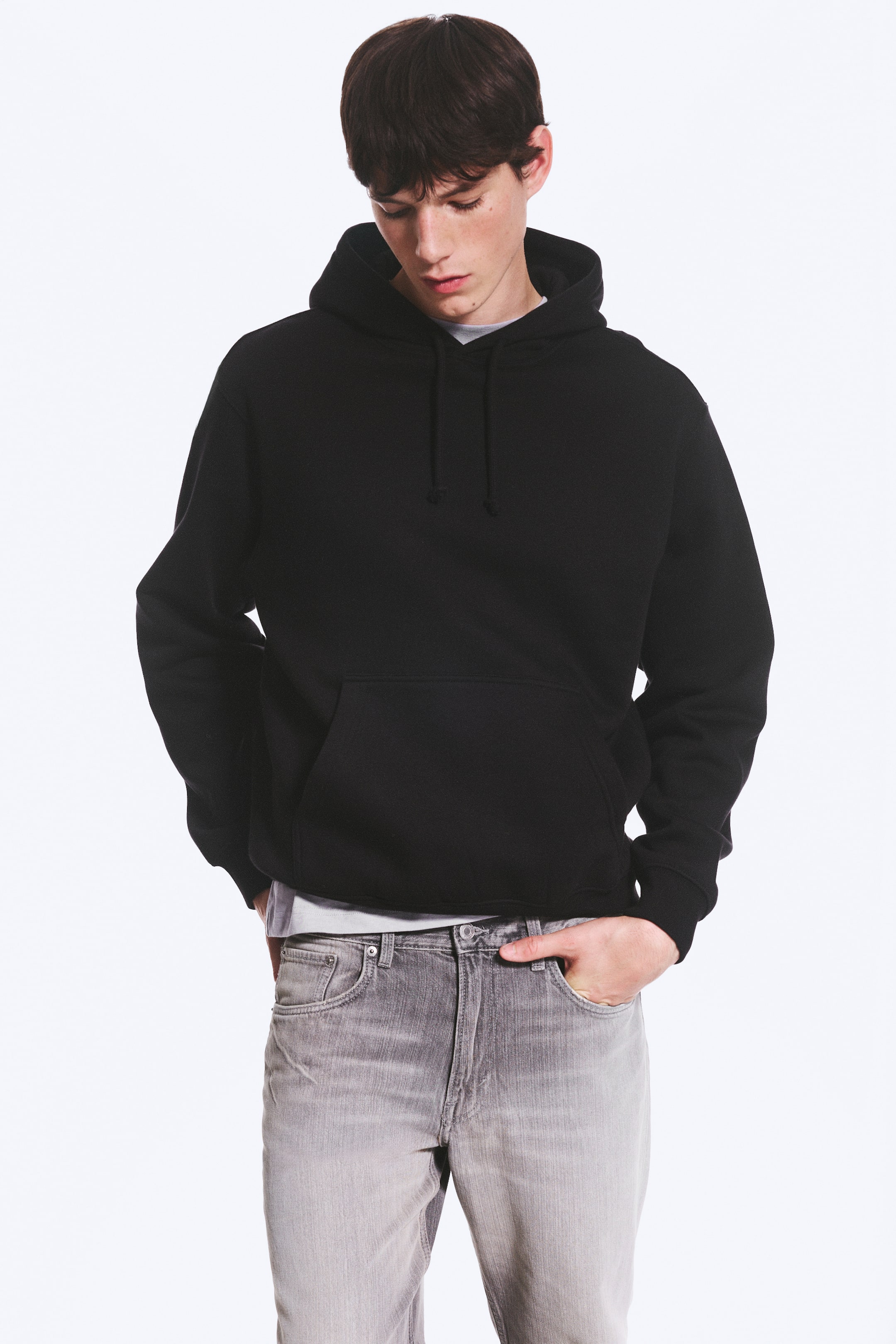 Visualizza immagine più grande: Una felpa nera con cappuccio viene presentata frontalmente, caratterizzata da un cappuccio con cordini neri, maniche lunghe con polsini a coste e una tasca a marsupio. Sotto viene indossata una t-shirt a girocollo di colore chiaro, con lo scollo e l'orlo che spuntano dal basso.