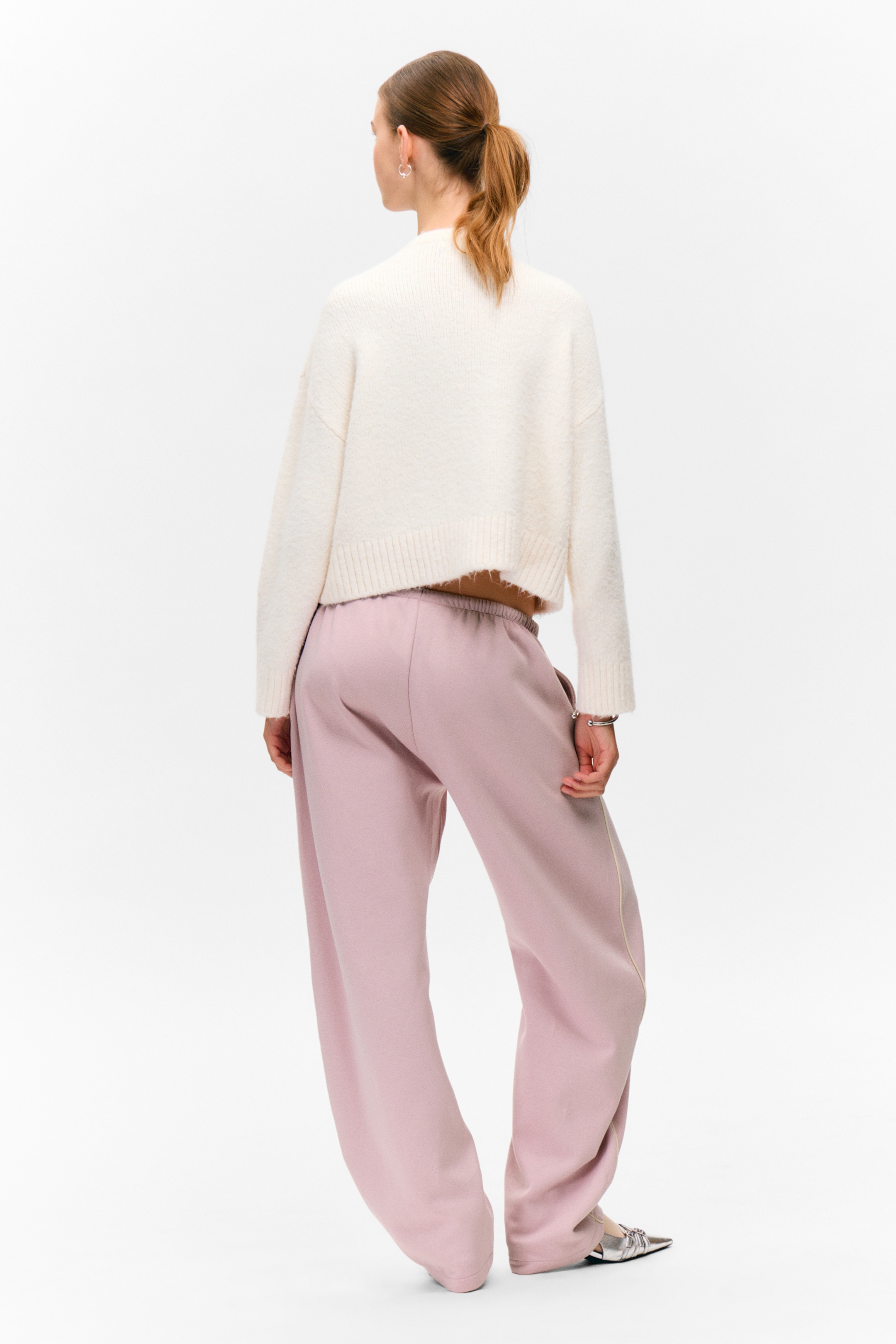 Ingrandisci l'immagine: Un maglione bianco a maglia tagliato, caratterizzato da una vestibilità rilassata e un orlo grezzo, è indossato sopra pantaloni rosa chiaro a gamba larga. I pantaloni sono progettati con una cintura elasticizzata e un sottile dettaglio a righe verticali lungo la cucitura laterale.
