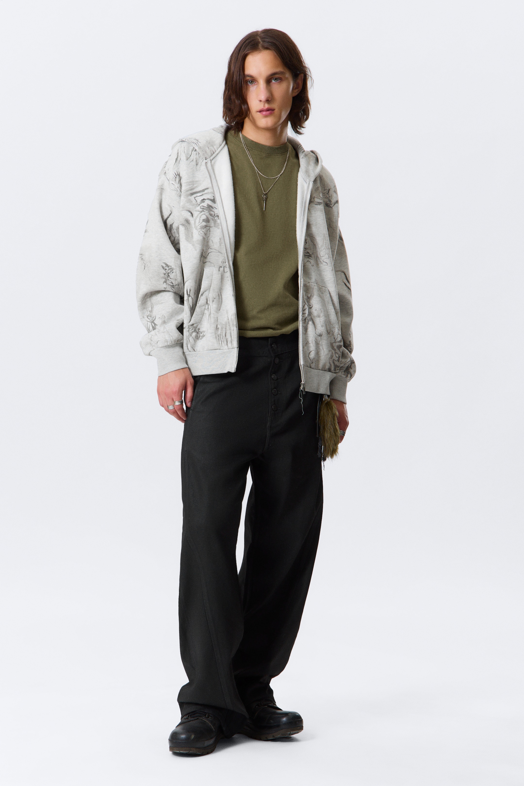 Visa större bild: En ljusgrå zip-up hoodie med ett mörkgrått grafiskt tryck över hela plagget bärs öppen, vilket avslöjar en olivgrön t-shirt med rund hals. Hoodien har ribbade muddar och fåll, medan svarta byxor med vida ben och en synlig knappgylf kompletterar den avslappnade silhuetten.