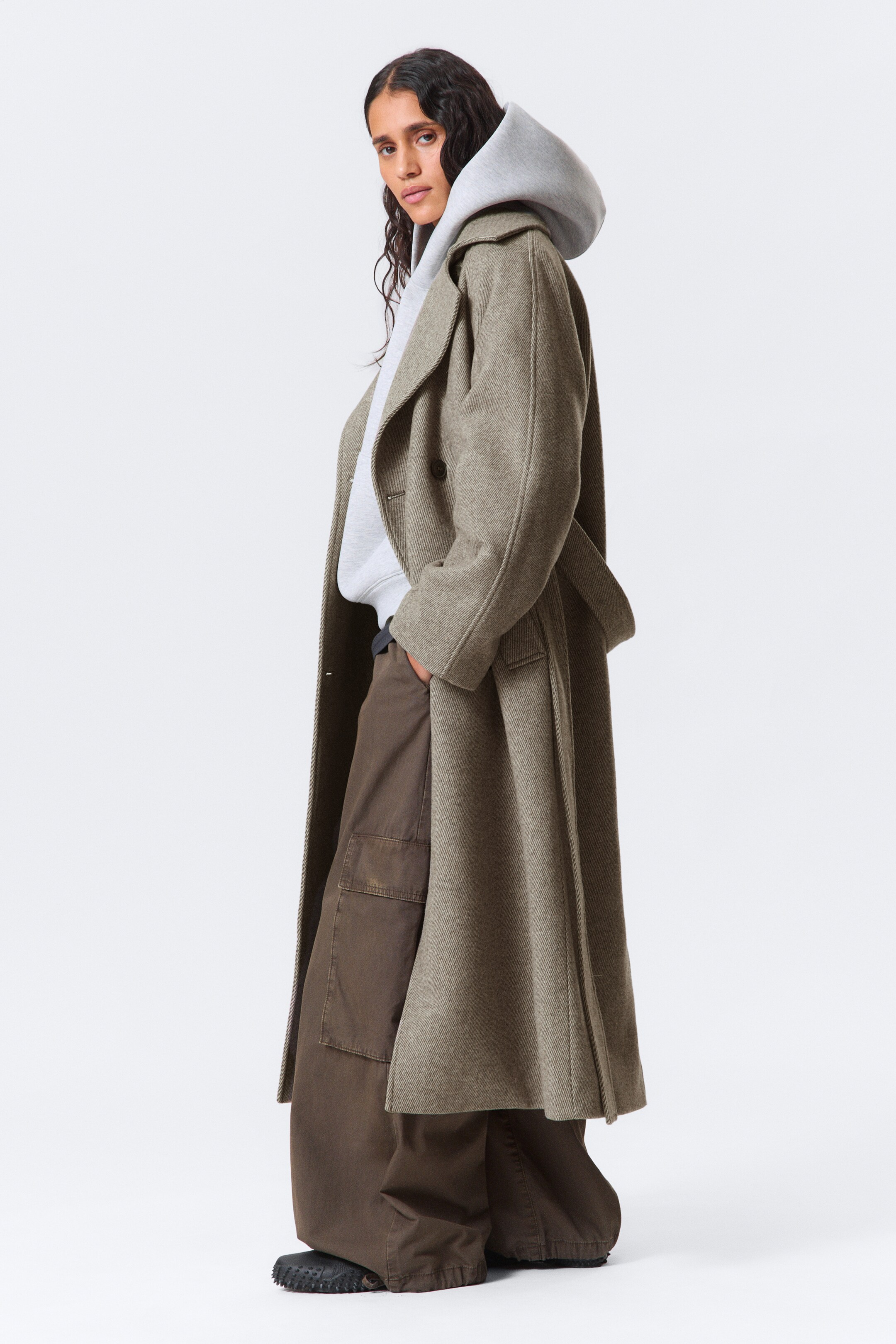 Agrandir l'image: Un long manteau surdimensionné à double boutonnage en mélange de laine texturée gris-vert est porté par le mannequin. Il présente de larges revers crantés et des manches raglan, avec une ceinture amovible drapée sur le côté, et le mannequin pose une main dans une poche passepoilée tout en superposant le manteau sur un sweat à capuche gris et un pantalon large marron.