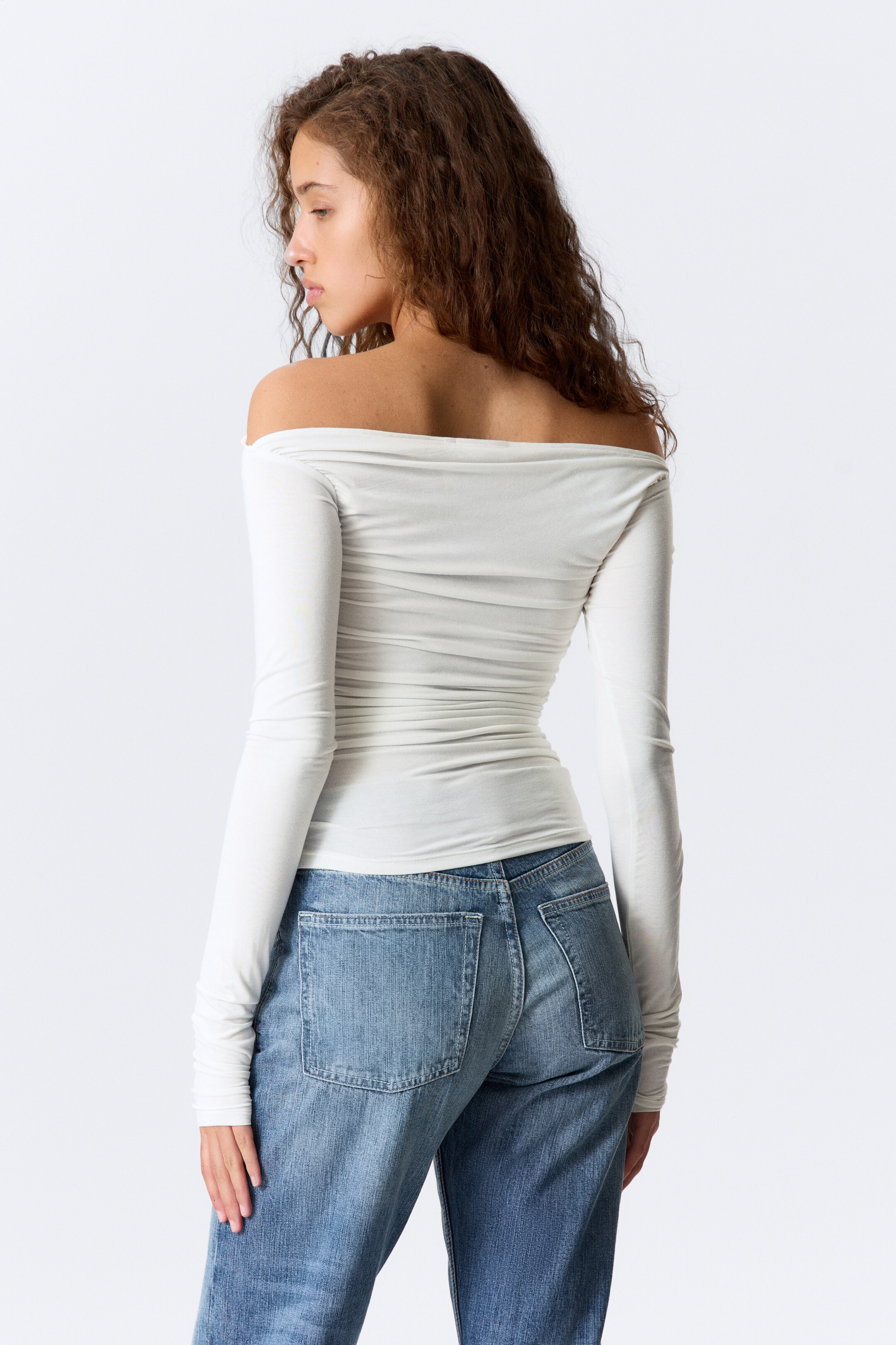 Agrandir l'image: Un haut blanc à manches longues est présenté de dos, légèrement incliné vers la gauche, avec un décolleté épaules dénudées, une silhouette ajustée et des fronces horizontales le long de son dos et de ses côtés.