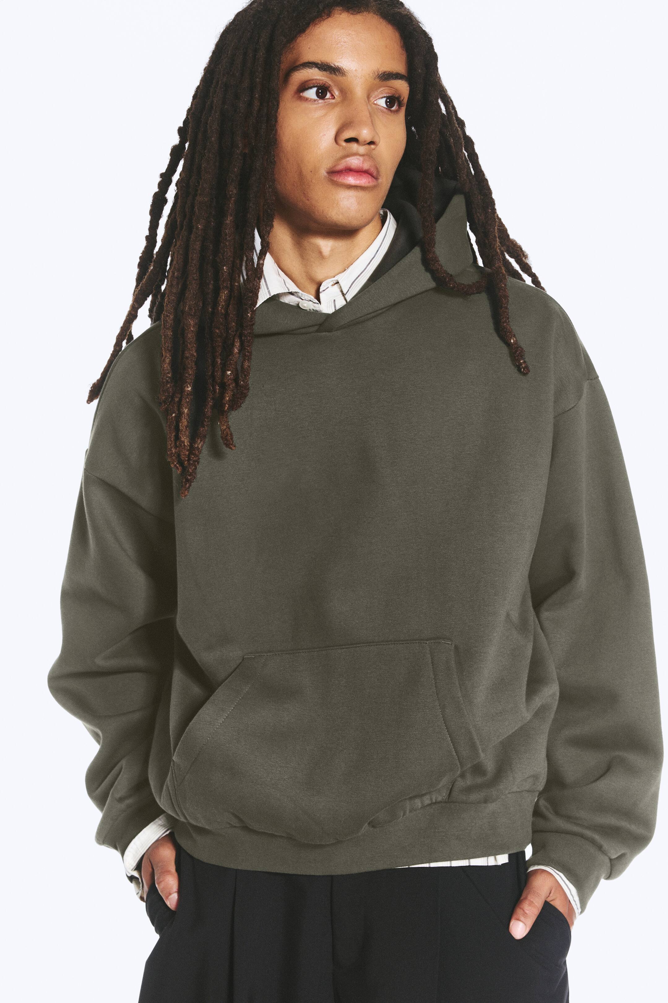 Grotere afbeelding bekijken: Een losvallende hoodie in donker olijfgroen, met afhangende schouders, een omgeslagen capuchonconstructie en een kangoeroezak aan de voorkant, wordt gedragen door het model. De hoodie is gelaagd over een gestreept hemd met kraag en gecombineerd met een donkere broek, waarbij het model zijn handen in de broekzakken steekt.