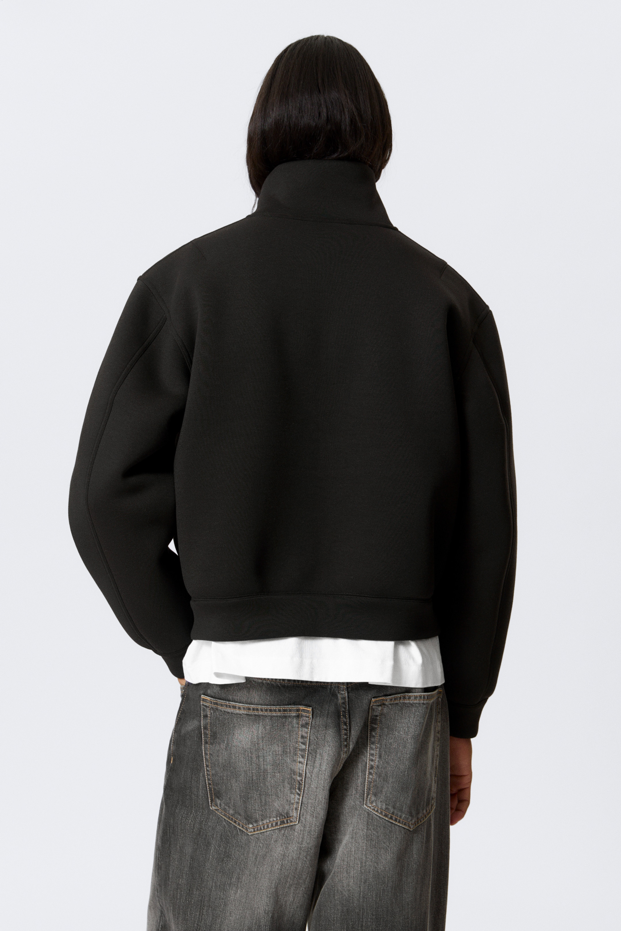 View larger image: A black jacket is being viewed from the back, featuring a high, standing collar and full, structured sleeves ending in fitted cuffs. A wide, fitted hem band is completing its cropped silhouette, with a white t-shirt peeking out from beneath.