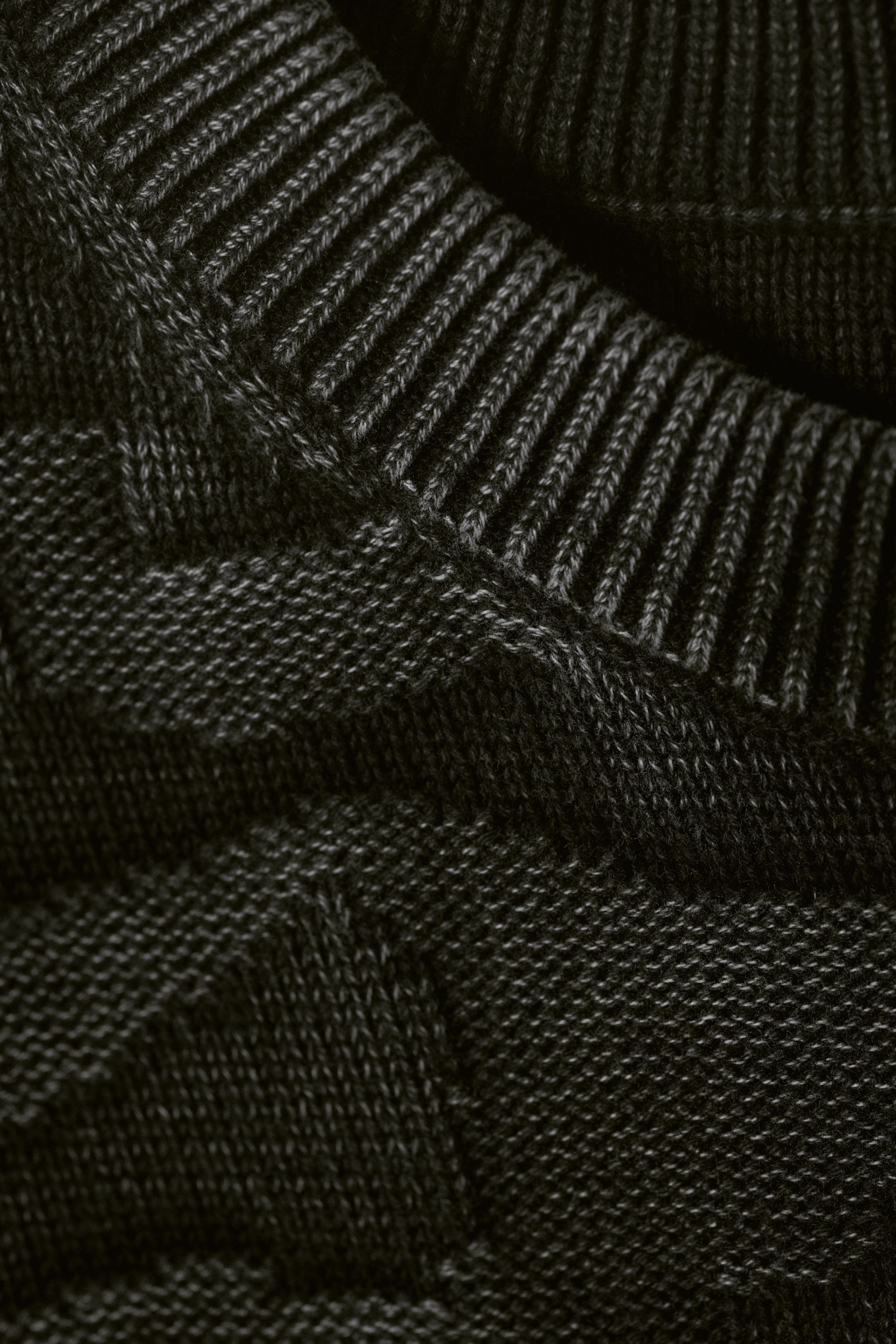 Größeres Bild anzeigen: Ein dunkles, meliertes Strickkleidungsstück wird in einer Nahaufnahme präsentiert, das einen gerippten Halsausschnitt und einen Hauptteil mit einem dezenten strukturierten Strickmuster zeigt.