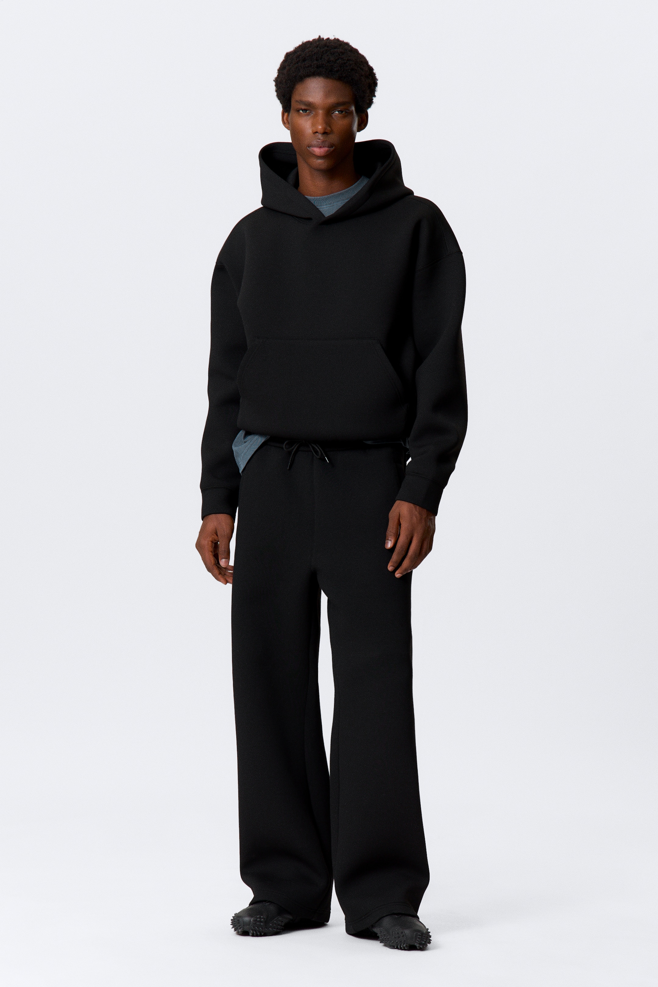 Agrandir l'image: Un sweat à capuche noir surdimensionné est présenté face avant, avec une capuche et une poche kangourou à l'avant, et est superposé à un t-shirt gris. Un pantalon noir à jambes larges assorti complète la tenue, les deux vêtements apparaissant dans un tissu tricoté texturé et le pantalon présentant une taille à cordon.