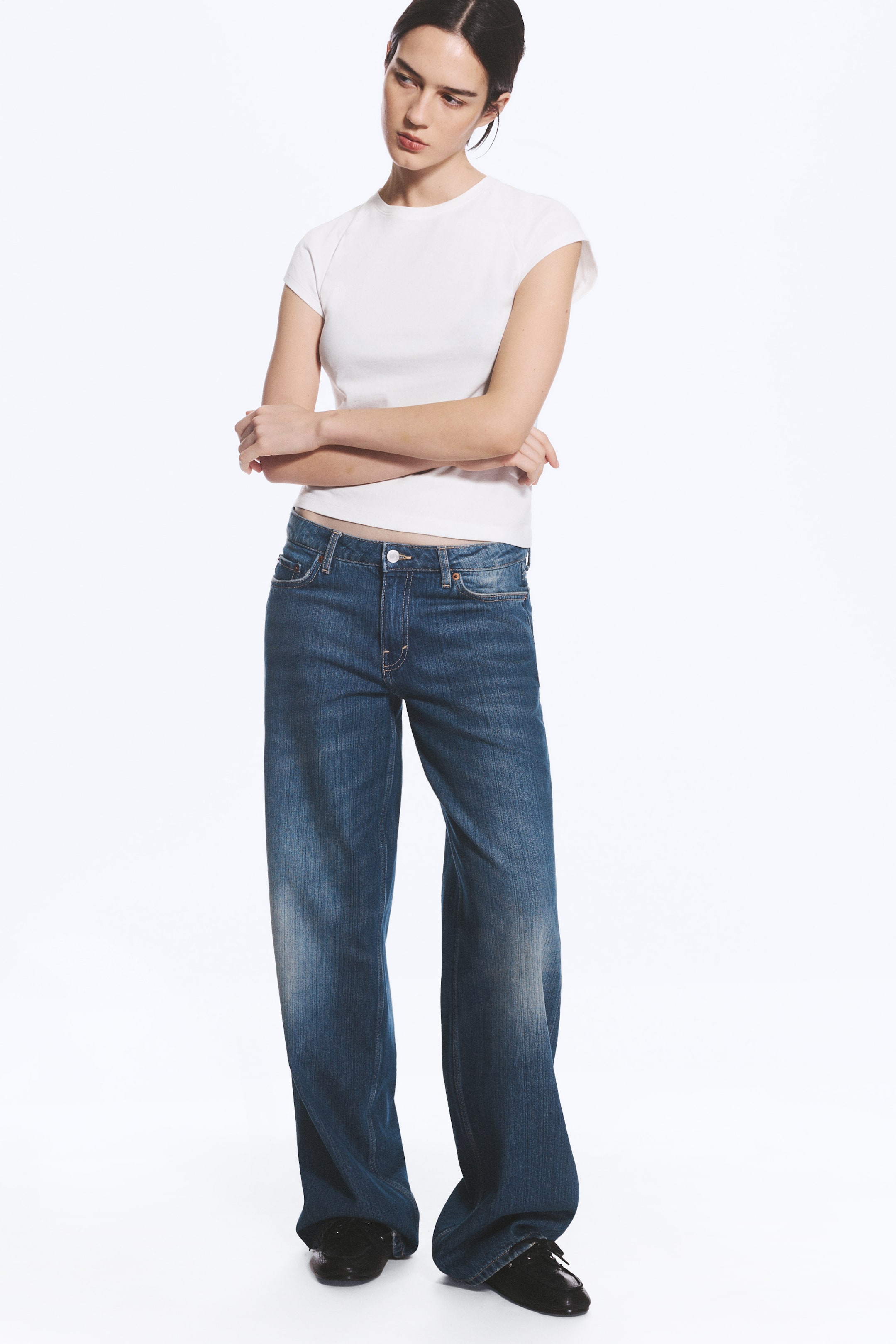 Agrandir l'image: Le mannequin porte un jean bleu taille basse à jambe large, à la coupe ample et à la longueur longue. Ce jean présente un délavage moyen avec de légères nuances délavées et est associé à un t-shirt blanc à manches courtes.