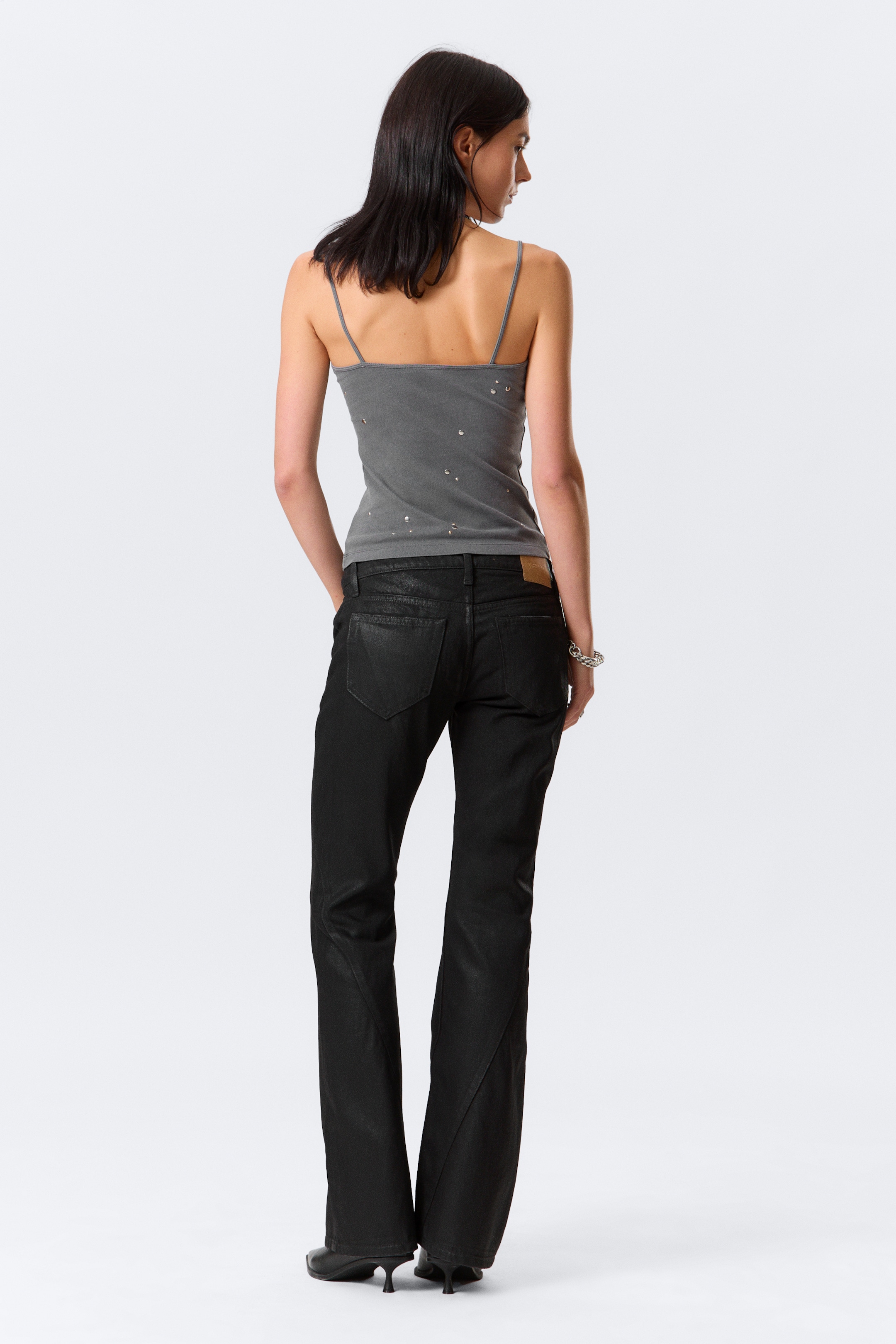 Agrandir l'image: Un débardeur gris, doté de fines bretelles spaghetti et de petites décorations brillantes éparses dans le dos, est présenté de dos. Le débardeur est porté avec un pantalon bootcut noir, légèrement brillant, qui présente un empiècement en cuir marron clair sur la ceinture arrière et des poches arrière simples.