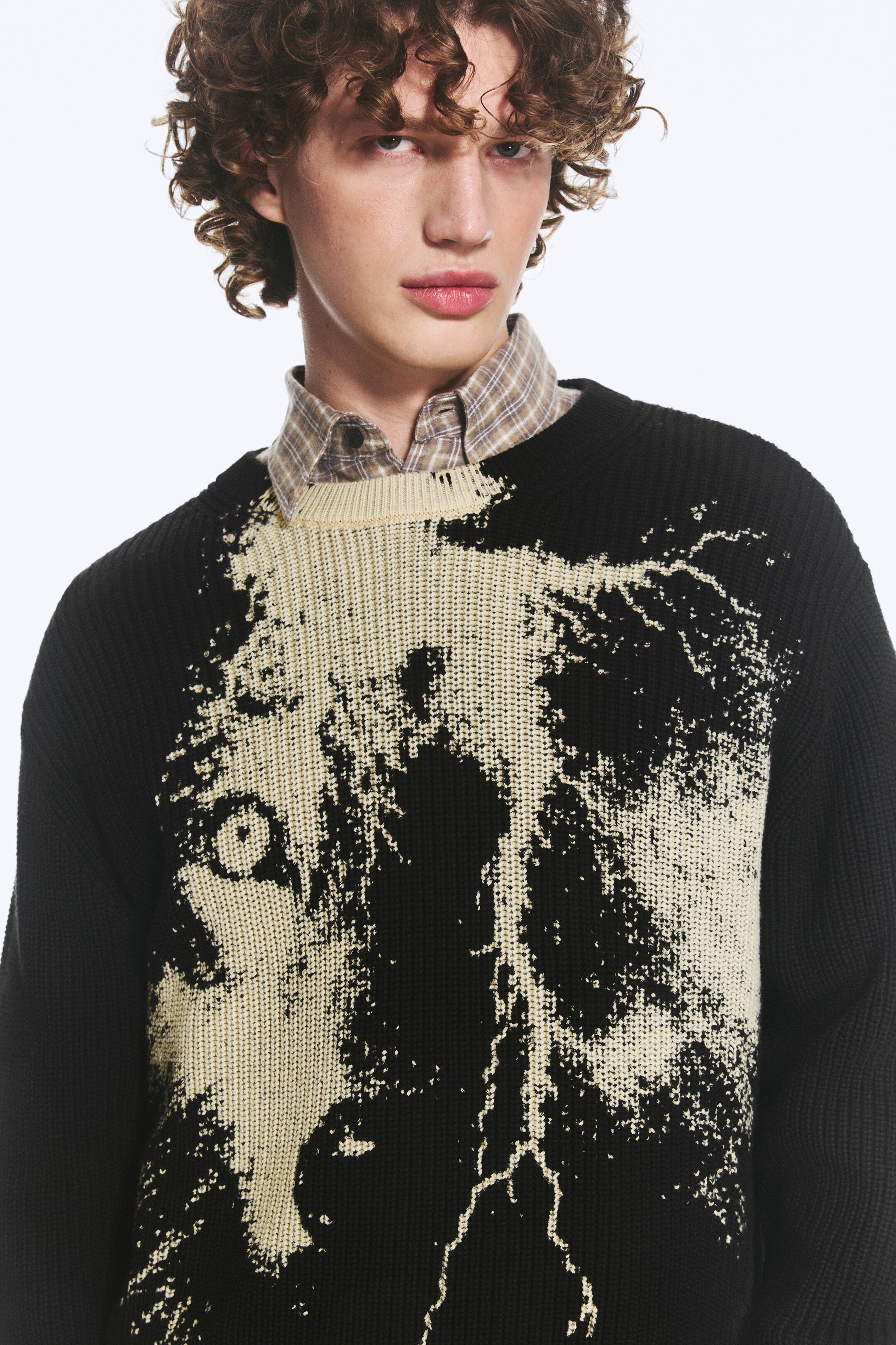 Ver imagen más grande: El jersey de punto negro muestra un gran gráfico abstracto de color crema en la parte frontal, posiblemente representando un rostro o una criatura estilizada. Presenta un cuello redondo acanalado y se lleva sobre una camisa de cuadros abotonada, con el cuello de la camisa visible.