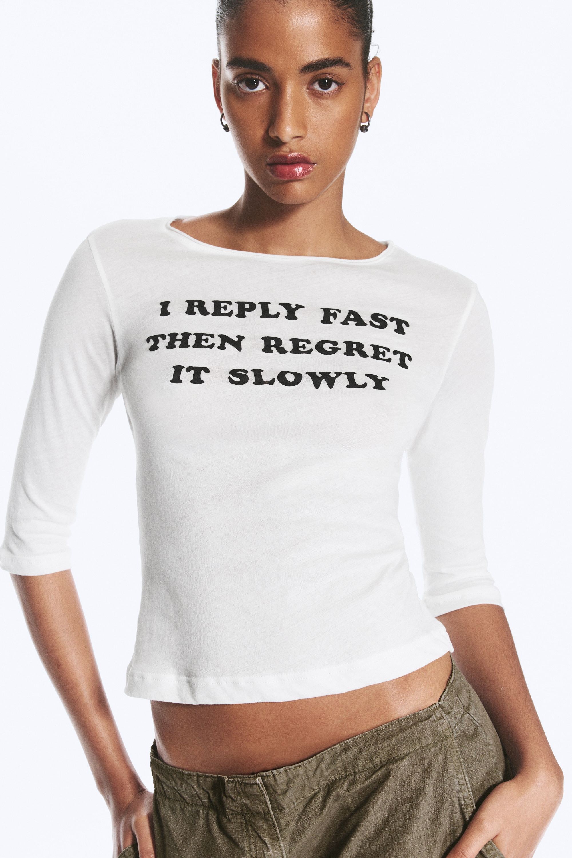 A white three-quarter-sleeved top is shown from the front, featuring a wide boat neckline and black text printed across the chest that reads, "I REPLY FAST THEN REGRET IT SLOWLY."