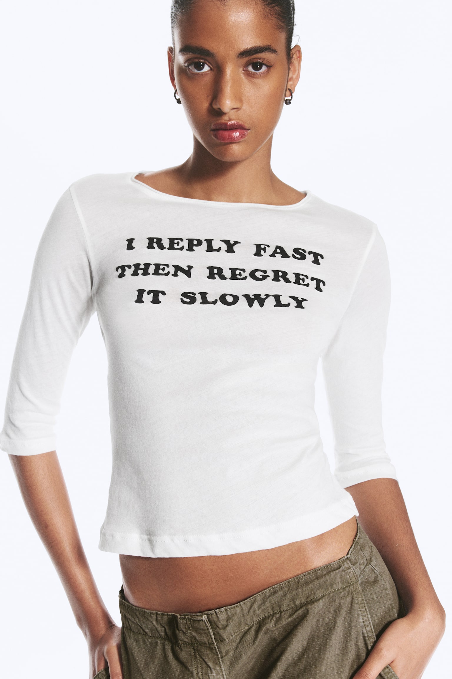 Uma camisola branca de manga três quartos está virada para a frente, exibindo um decote barco largo e texto preto impresso no peito com a frase: "I REPLY FAST THEN REGRET IT SLOWLY." - 1