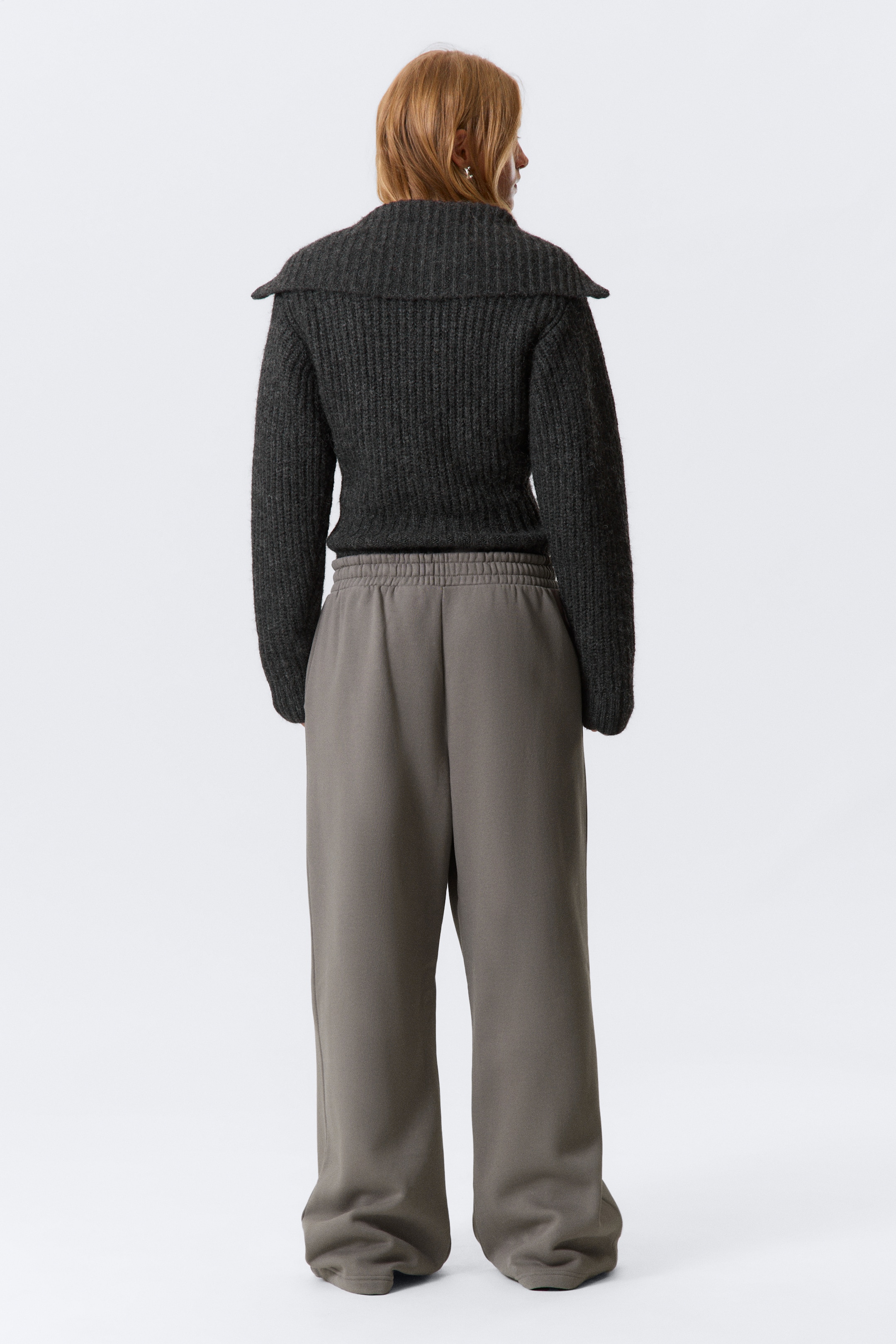 Visa större bild: En mörkgrå ribbstickad tröja visas från baksidan, med en bred sjalkrage och en något kortare fåll. Den bärs med löst sittande, vida gråa sweatpants som samlas vid anklarna och har en elastisk midja.