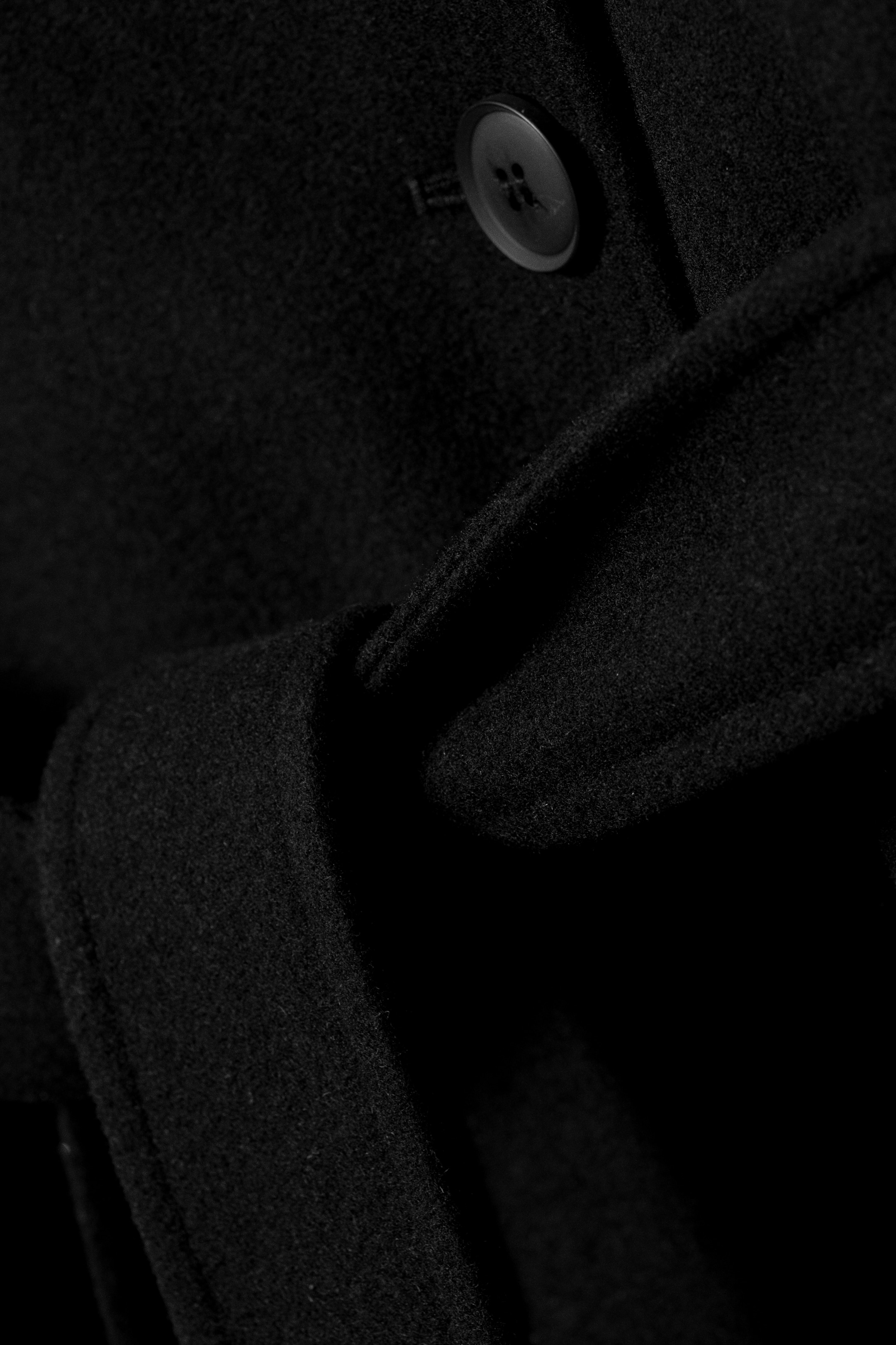 Visualizza immagine più grande: Un cappotto in misto lana nero mostra la sua finitura spazzolata, con un bottone tonale visibile sulla paramontura. Un cinturino coordinato stringe il tessuto, creando morbide pieghe.