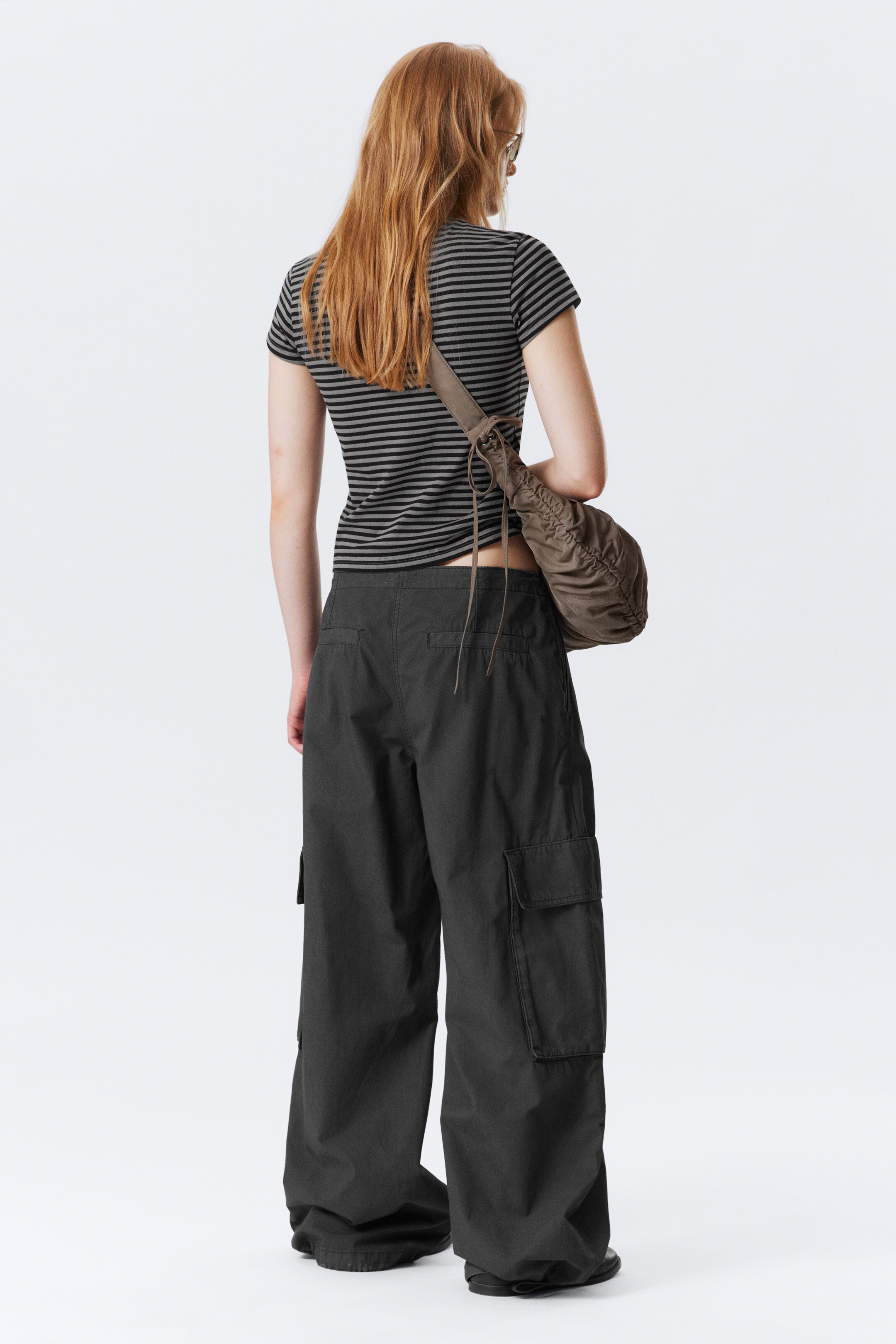 Vezi imaginea mai mare: Un tricou cu mâneci scurte, cu dungi gri închise și deschise, este purtat cu pantaloni cargo largi și închiși la culoare, care au buzunare mari aplicate pe părțile laterale ale picioarelor. O geantă de umăr maro, strânsă, este purtată pe spatele ținutei.