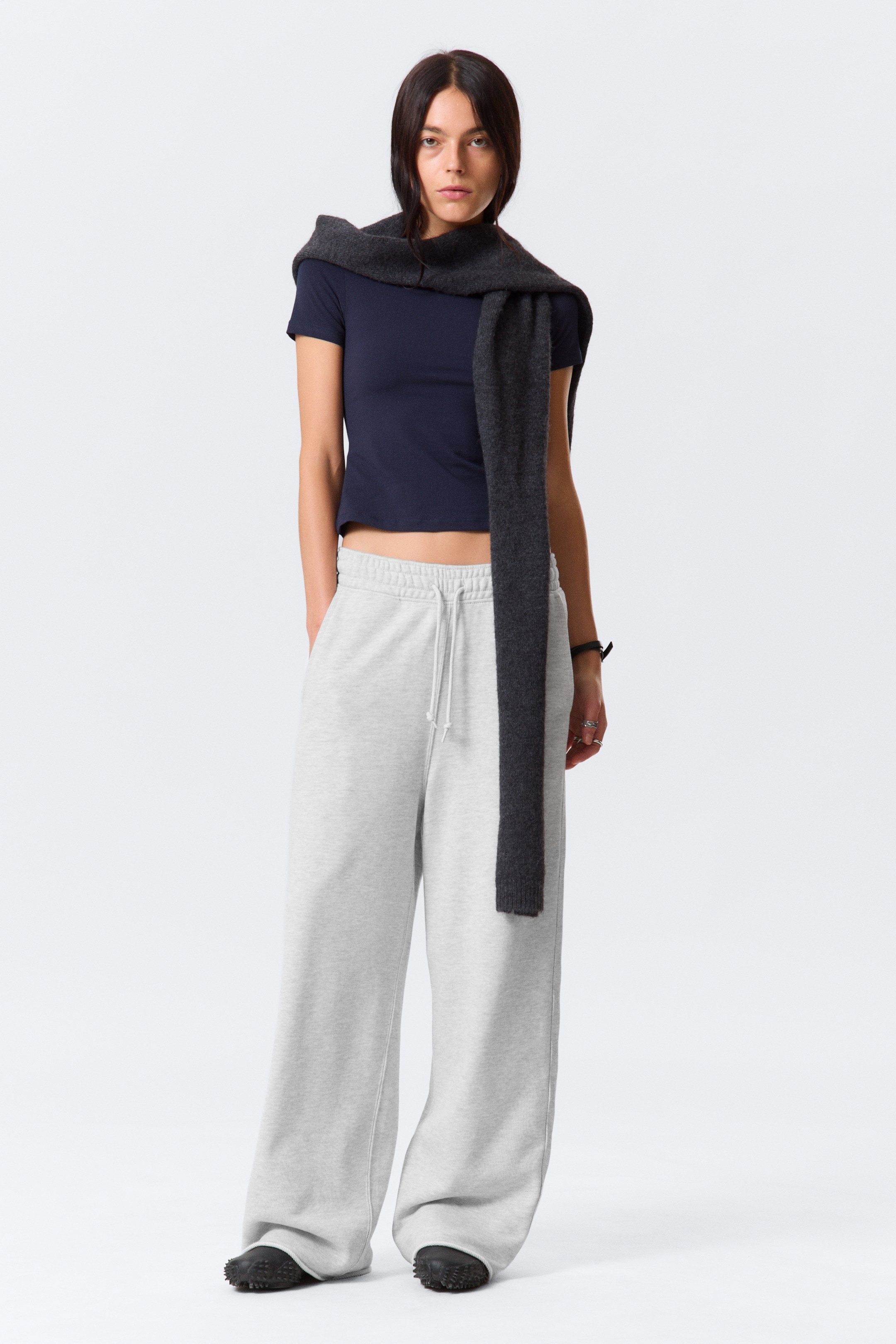 Agrandir l'image: Un pantalon de survêtement gris clair, ample et à jambes larges est porté par le mannequin. Ils sont fabriqués en éponge de coton épaisse et présentent une large ceinture à cordon de serrage, des poches latérales en biais, et des ourlets non traités. Le mannequin se tient debout avec les mains dans les poches latérales en biais.