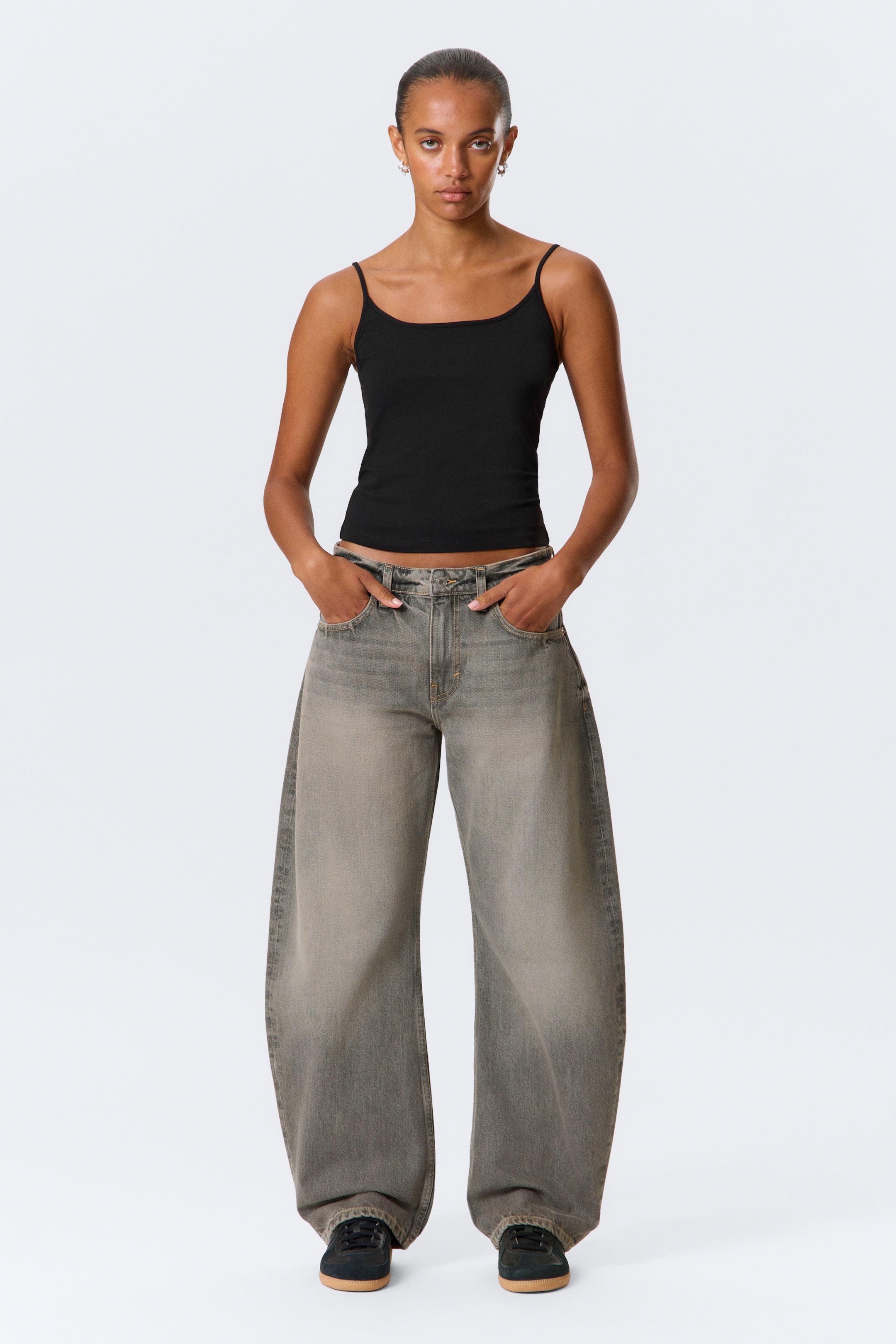 Grösseres Bild ansehen: Ein schwarzes geripptes Camisole-Tanktop wird getragen, das über dünne Spaghettiträger und einen Rundhalsausschnitt verfügt. Es wird mit ausgewaschenen grauen, tief sitzenden Jeans mit weitem Bein kombiniert, die mit einer lockeren Passform gestaltet sind und an den Knöcheln zusammenfallen.