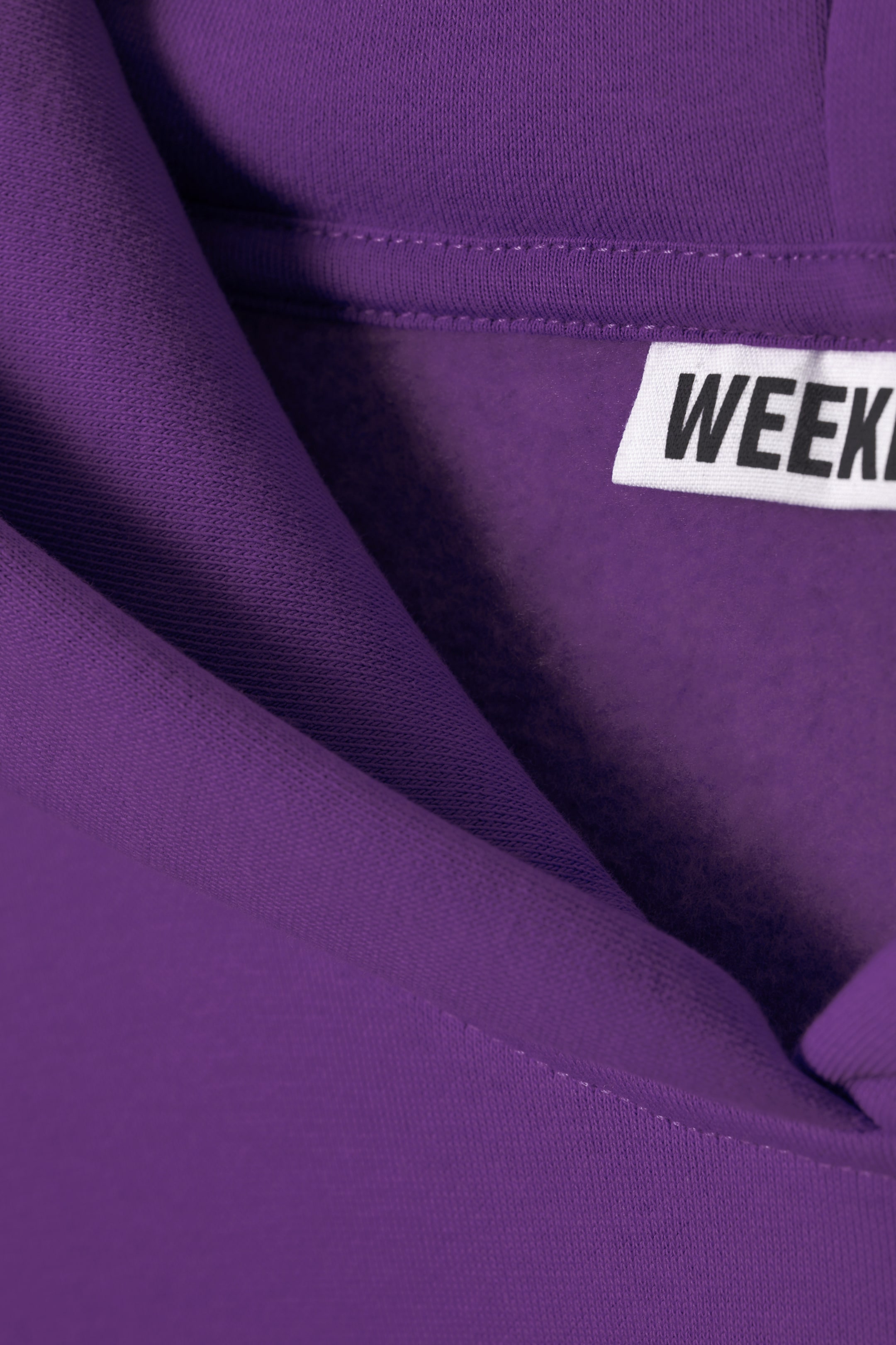Grotere afbeelding bekijken: Een close-up van een hoekige kijk presenteert een paarse kledingstuk, waarbij de gebreide buitenstof en zachtere binnenstof worden getoond. Een wit label, waarop gedeeltelijk 'WEEK' te lezen is, is zichtbaar aan de binnenkant van de gevouwen rand van het kledingstuk, waar een dubbelgestikte naad langs de opening loopt.