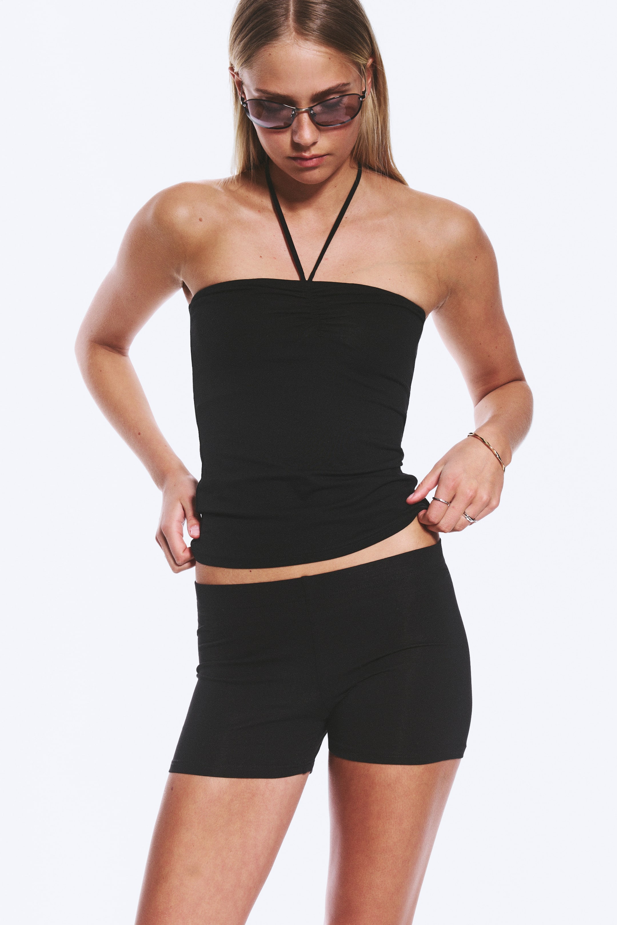 Größeres Bild ansehen: Das Model trägt ein schwarzes Neckholder-Top und schwarze, eng anliegende Shorts. Das Neckholder-Top hat eine geraffte Brustpartie, und das Model passt mit beiden Händen den unteren Saum an.