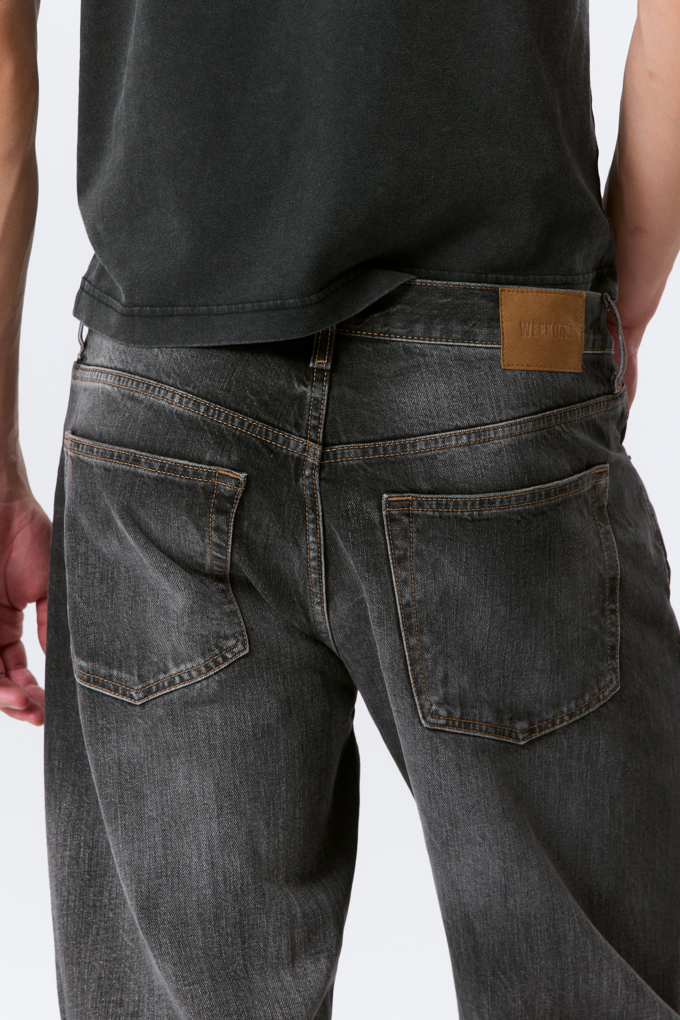 Größeres Bild ansehen: Dunkelgraue Waschjeans werden von hinten gezeigt, mit hellbraunen Kontrastnähten, die die beiden hinteren aufgesetzten Taschen und den Bund umreißen. Ein hellbraunes rechteckiges Markenetikett ist auf der rechten Seite des hinteren Bunds sichtbar.