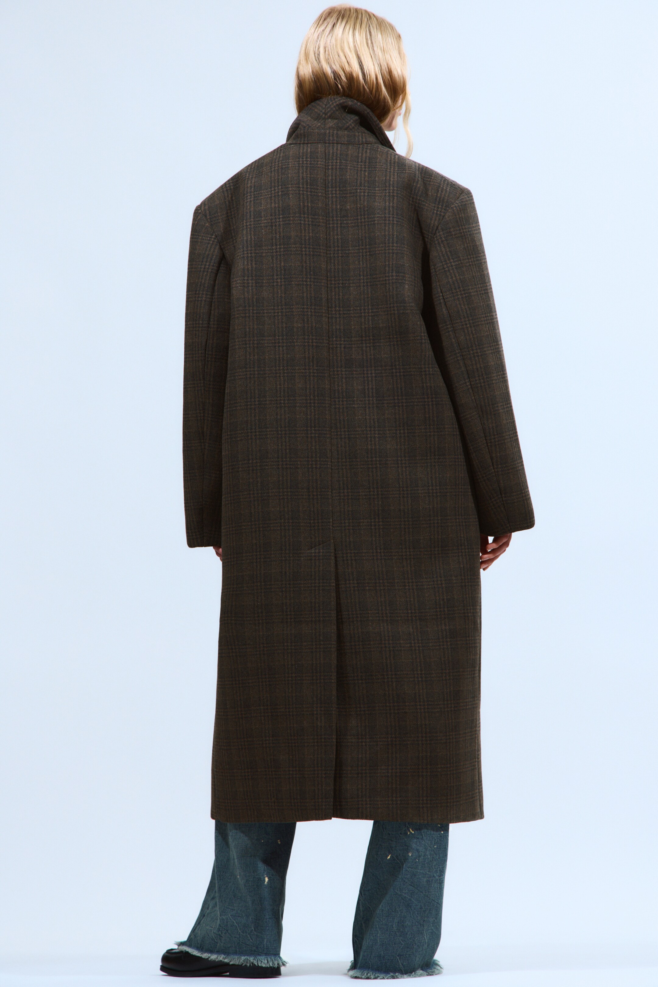 View larger image: A dark brown checked coat is being worn, viewed from the back. The long, relaxed-fit coat features a visible collar and a back vent, extending below the knees over blue jeans.